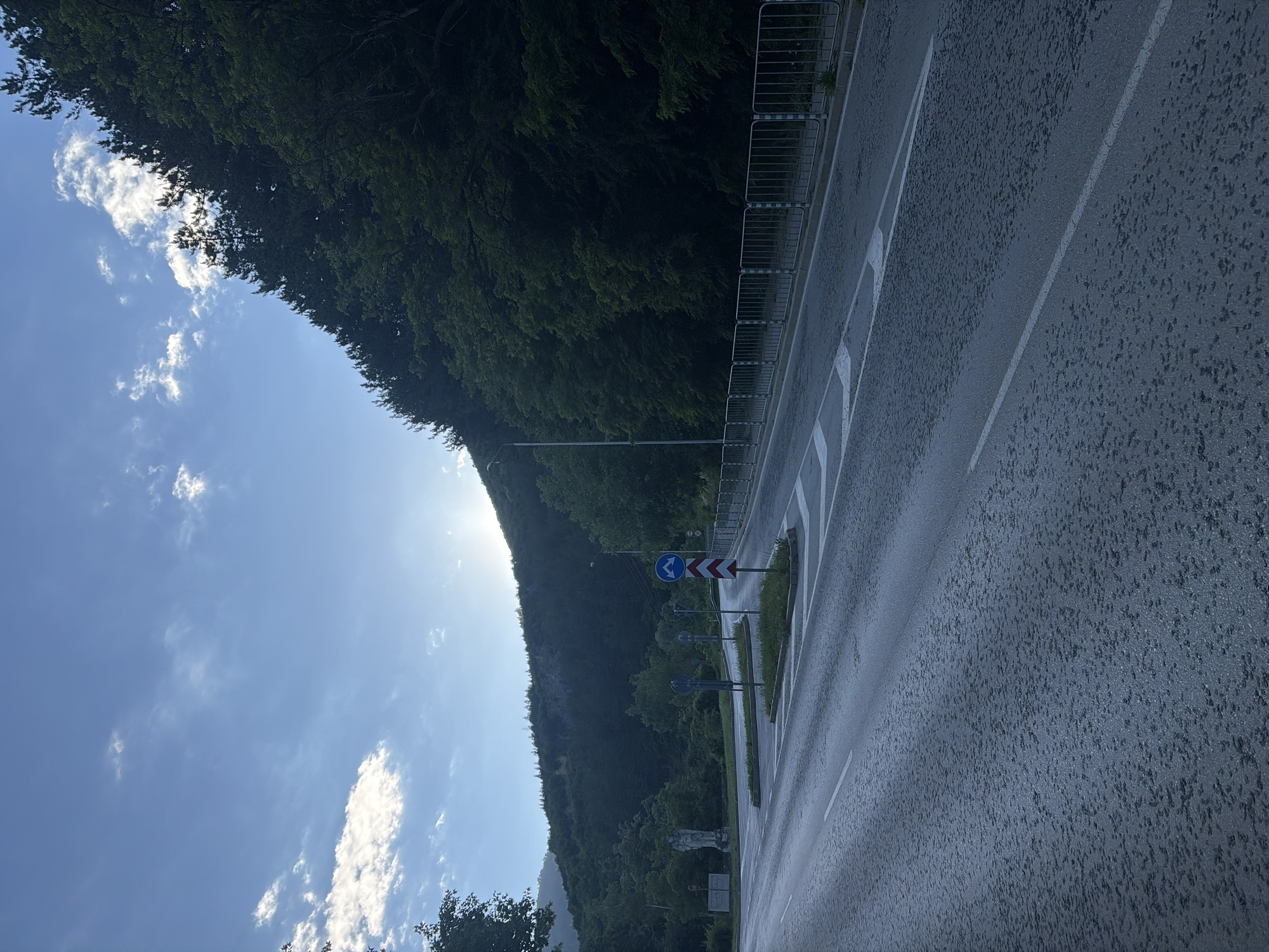 The image depicts a road junction in Vratsa, Bulgaria, with a mountain range covered in dense trees forming the backdrop. The sky is blue with scattered white clouds, and the sun is setting or rising, casting a warm glow. The road appears wet, possibly from recent rain, and has lane markings. There are several road signs, including directional signs and traffic warnings. A metal barrier runs along the side of the road, separating it from the wooded area. In the distance, a statue is visible on the left side of the road. The scene is quiet and serene, with no people or vehicles present.