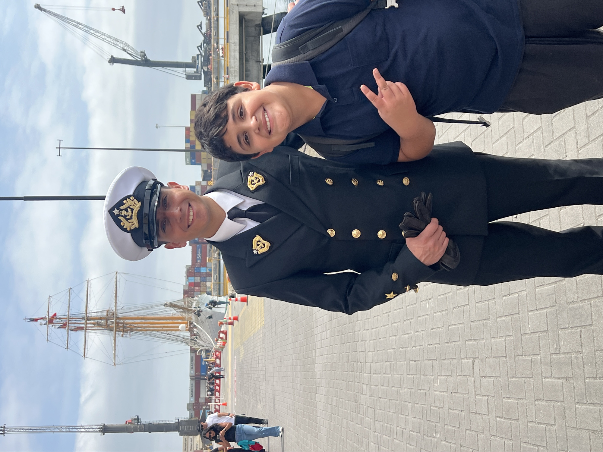 Arica, Chile: A man in a naval uniform stands with a boy on a paved area near a port. The man is wearing a white naval cap, a dark blue uniform with gold buttons, a black tie, and black gloves. The boy, with a backpack, wears a dark blue collared shirt and black pants, giving a peace sign. In the background are shipping containers, cranes, a sailing ship, and other people. The sky is partially cloudy.