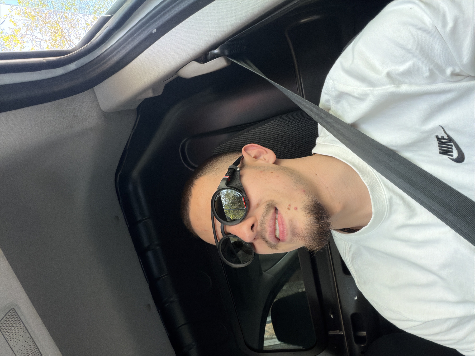 The image, taken in Plovdiv, Bulgaria, features a young man seated inside a vehicle during daylight hours. He is seen from the chest up, wearing a white short-sleeved t-shirt that prominently displays a black "NIKE" logo on the right side of his chest. He also wears dark, round sunglasses; the brand "Carrera" is faintly discernible on the frame of the sunglasses. A black seatbelt is fastened across his chest, extending diagonally from his right shoulder.
The man has a beard or stubble and looks towards the viewer with a slight smile, indicating a pleasant or relaxed demeanor. The reflections in his sunglasses reveal bright green and yellow foliage against a clear sky, consistent with sunny weather. The background primarily consists of the vehicle's interior, including a white headliner, dark gray and black plastic trim, and a window through which more trees and sky are visible. A texture resembling a speaker grill is visible on an interior panel in one of the cropped views. The image is notably tilted, suggesting an unusual camera angle or the subject leaning back. The bright natural light indicates it is daytime.