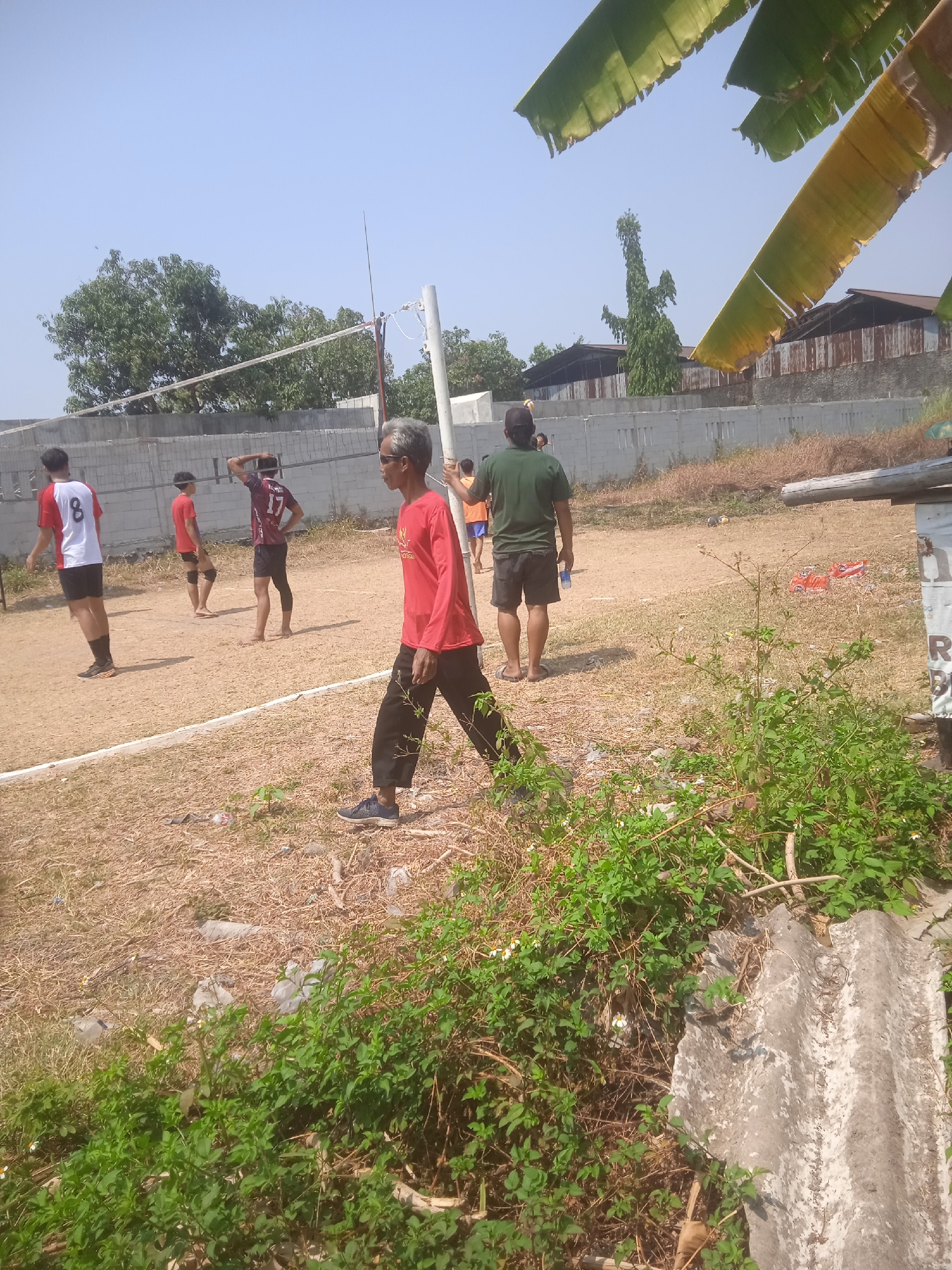 In Klodran, Indonesia, a group of individuals are engaged in a game of volleyball on a dirt court. The scene depicts several people, predominantly younger men wearing athletic attire, and at least one older man with gray hair, also dressed in sportswear. The court is marked with white lines and features a volleyball net supported by poles. The surrounding environment is dry and somewhat arid, with sparse vegetation and a backdrop of simple buildings, possibly housing or industrial structures, and a clear, bright sky. The lighting suggests it is daytime, with the sun casting shadows on the ground.