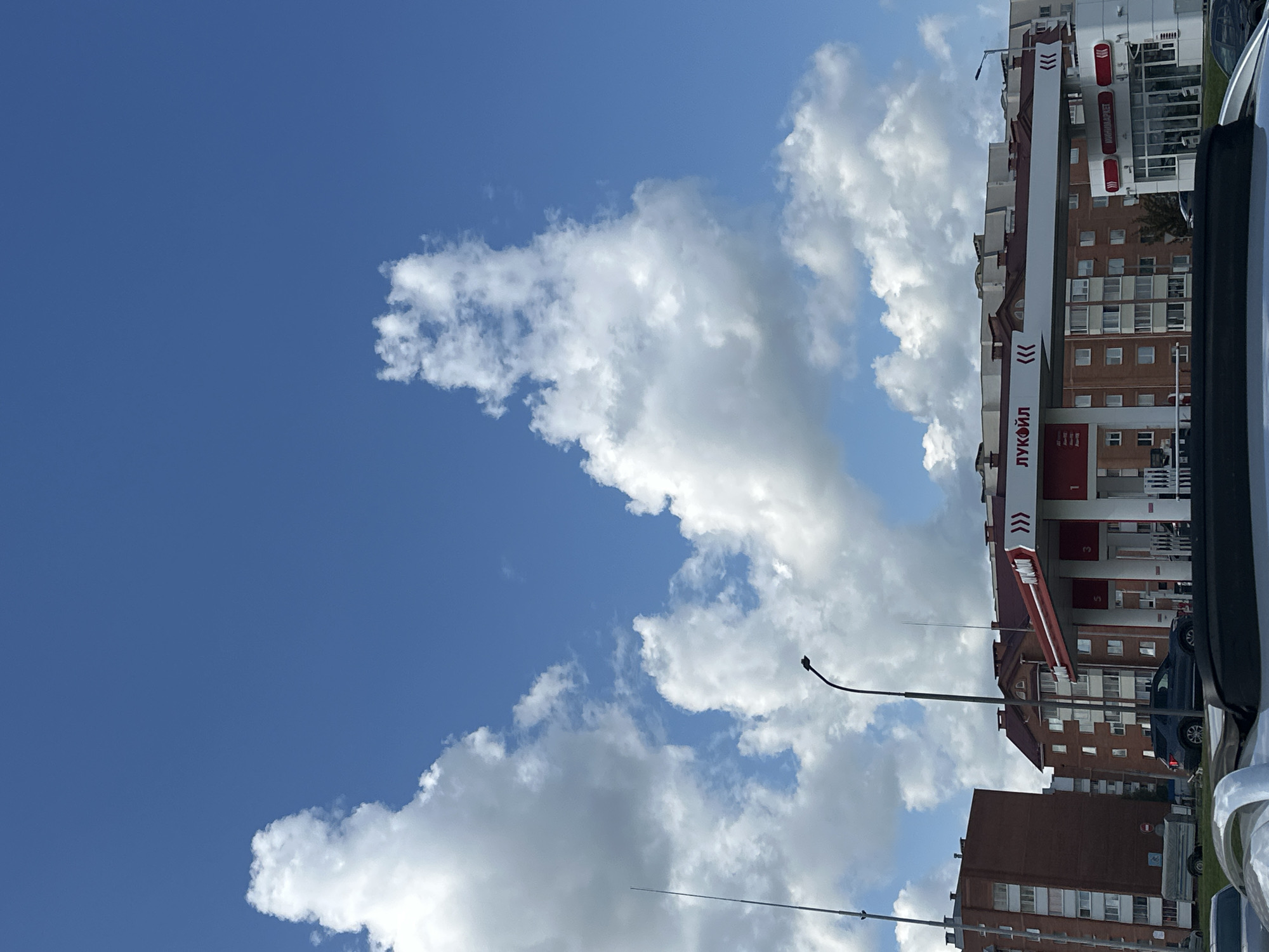 The image shows a view of a Lukoil gas station in Maladzyechna, Belarus, under a partly cloudy blue sky. In the foreground, the roof of a vehicle is visible. The gas station has a red and white canopy with the Lukoil logo. There are several cars parked at the station, including a dark SUV. Behind the gas station are multi-story brick buildings with red-tiled roofs. A street light is visible to the left of the gas station. The overall scene depicts a typical urban environment on a sunny day.