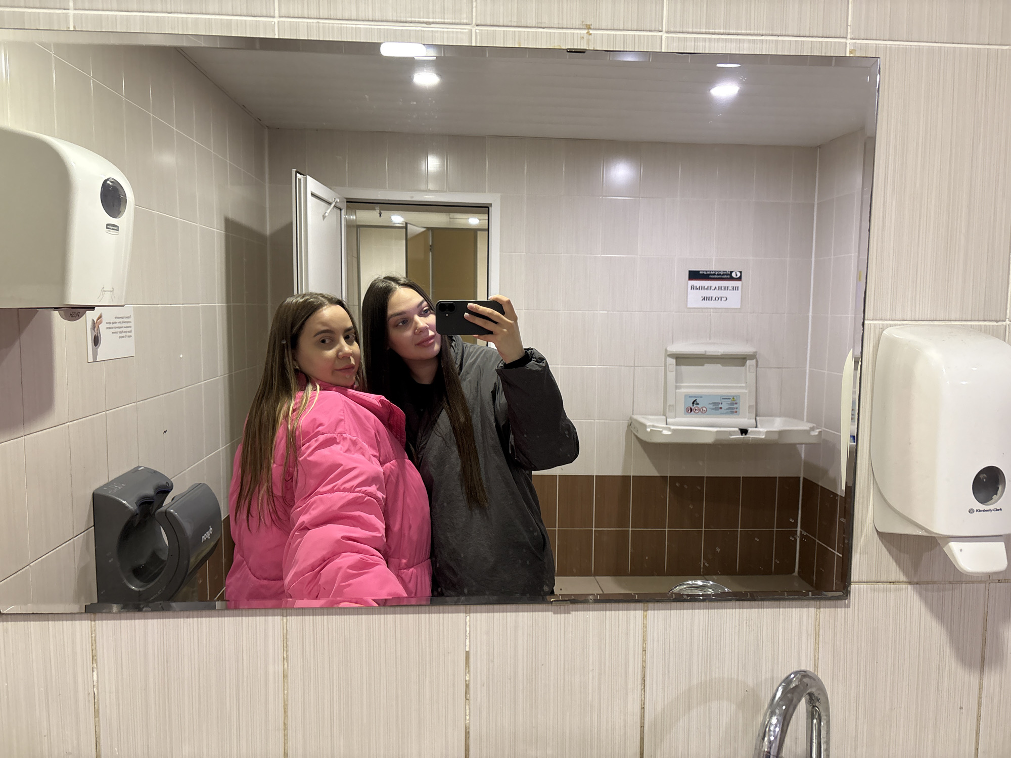 The image shows a mirror selfie taken in a public restroom, likely in Izhevsk, Russia, based on signage visible in the reflection. Two young women are visible in the mirror. The woman on the left wears a bright pink puffer jacket. The woman on the right is holding a cell phone and wearing a dark grey coat. The restroom features light-colored tiled walls with darker brown accents. Visible fixtures include a paper towel dispenser, a hand dryer, a baby changing station, a sink and faucet. A doorway is visible in the reflection, suggesting another room or area of the restroom.