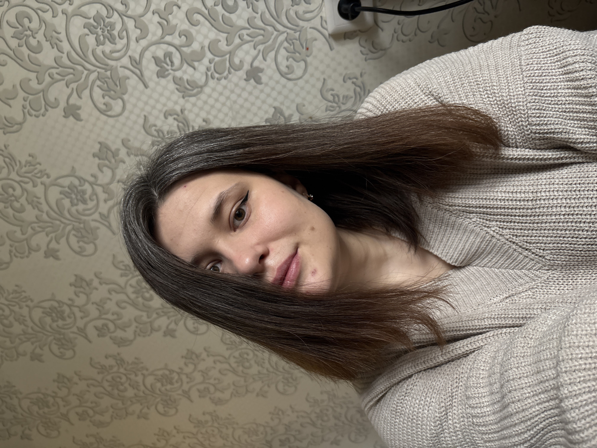 A woman is shown from the chest up, turned slightly towards the viewer and leaning towards the left of the frame. She has light skin, brown eyes, and brown hair with grey roots. She is wearing eyeliner on her upper eyelids. She is wearing a beige sweater. The background is a wall covered in a beige and white patterned wallpaper. A white electrical outlet with a black plug is visible at the top center. The location is Chaikovsky, Russia.