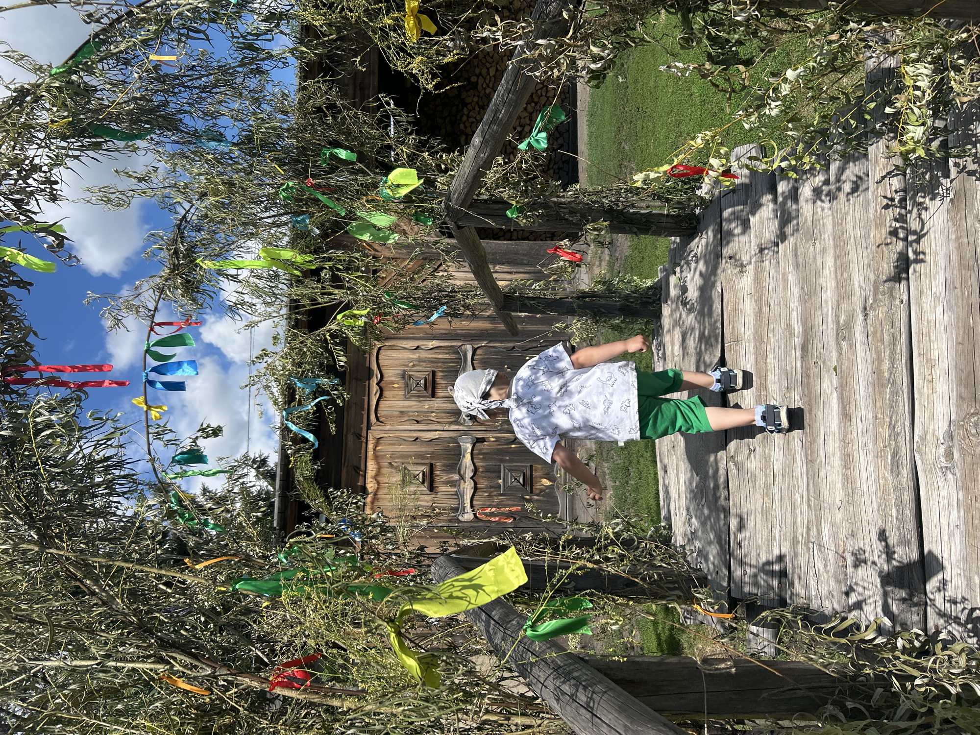 In Bolsherechye, Russia, a child wearing a white bandana, a white t-shirt with a dinosaur print, and green shorts walks across a wooden bridge towards a rustic wooden building with large double doors. The bridge and the entrance to the building are adorned with hanging green, red, yellow, and blue ribbons, as well as branches from a tree with long, wispy leaves. The scene is illuminated by natural daylight, with a blue sky and white clouds visible above. The ground beyond the bridge is covered in green grass, and to the right of the building, a stack of firewood is visible. The overall atmosphere suggests a festive or celebratory setting, possibly a village event or a park.