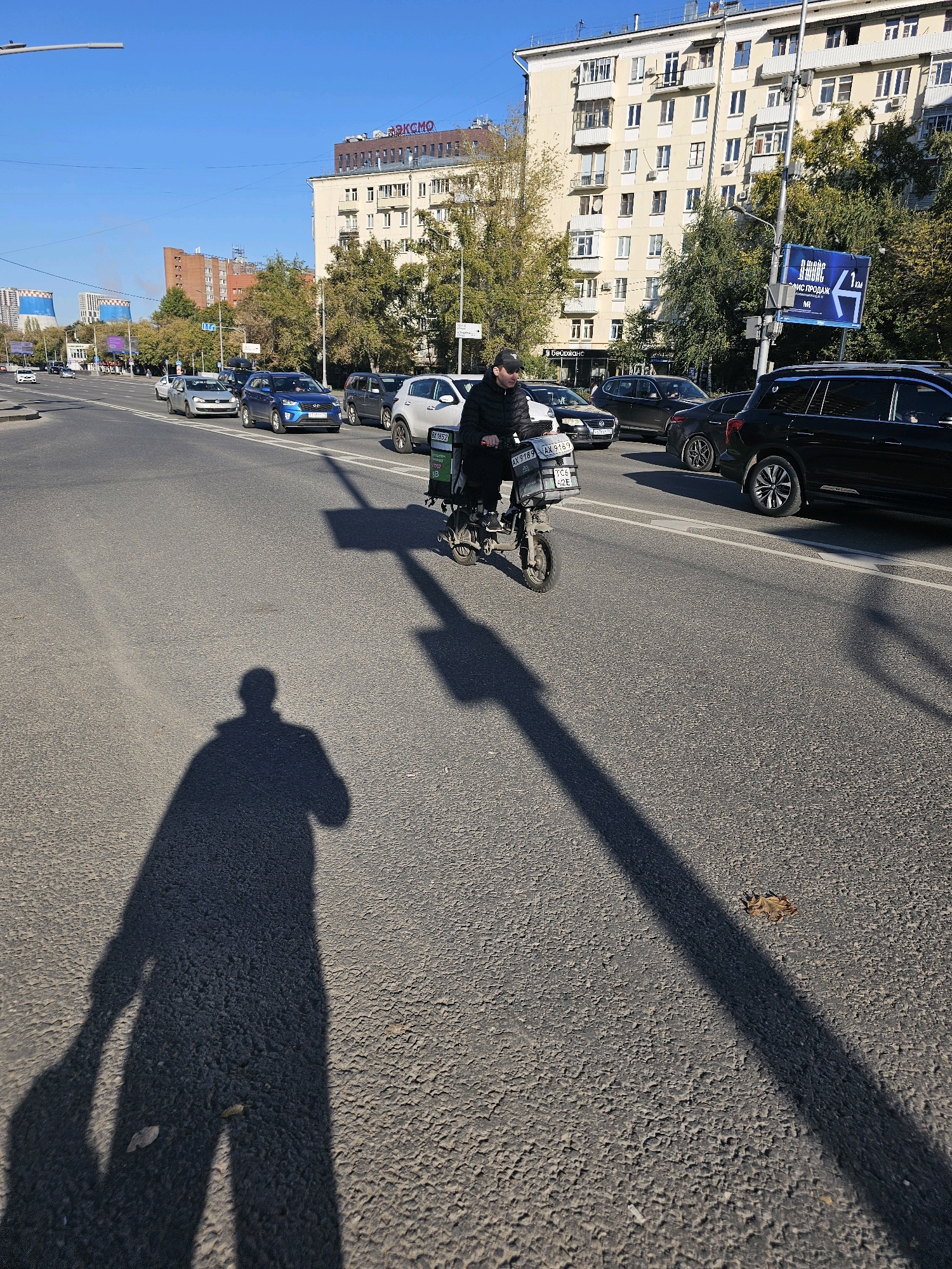 In Moscow, Russia, a person is riding an electric scooter with a delivery box on the back along a multi-lane road. The scooter has a license plate that reads "AX 9189 AX 9189" and "TCS 612 E". The rider is wearing a black cap and a dark jacket. The road is asphalt and appears to be well-maintained. In the background, there are several cars, buildings, and trees. The sky is clear and blue, suggesting it is a sunny day. A long shadow of the scooter rider and another person, who is not visible in frame but is likely the photographer, are cast on the road. A billboard with a map and text in Russian is visible on the right side of the image.