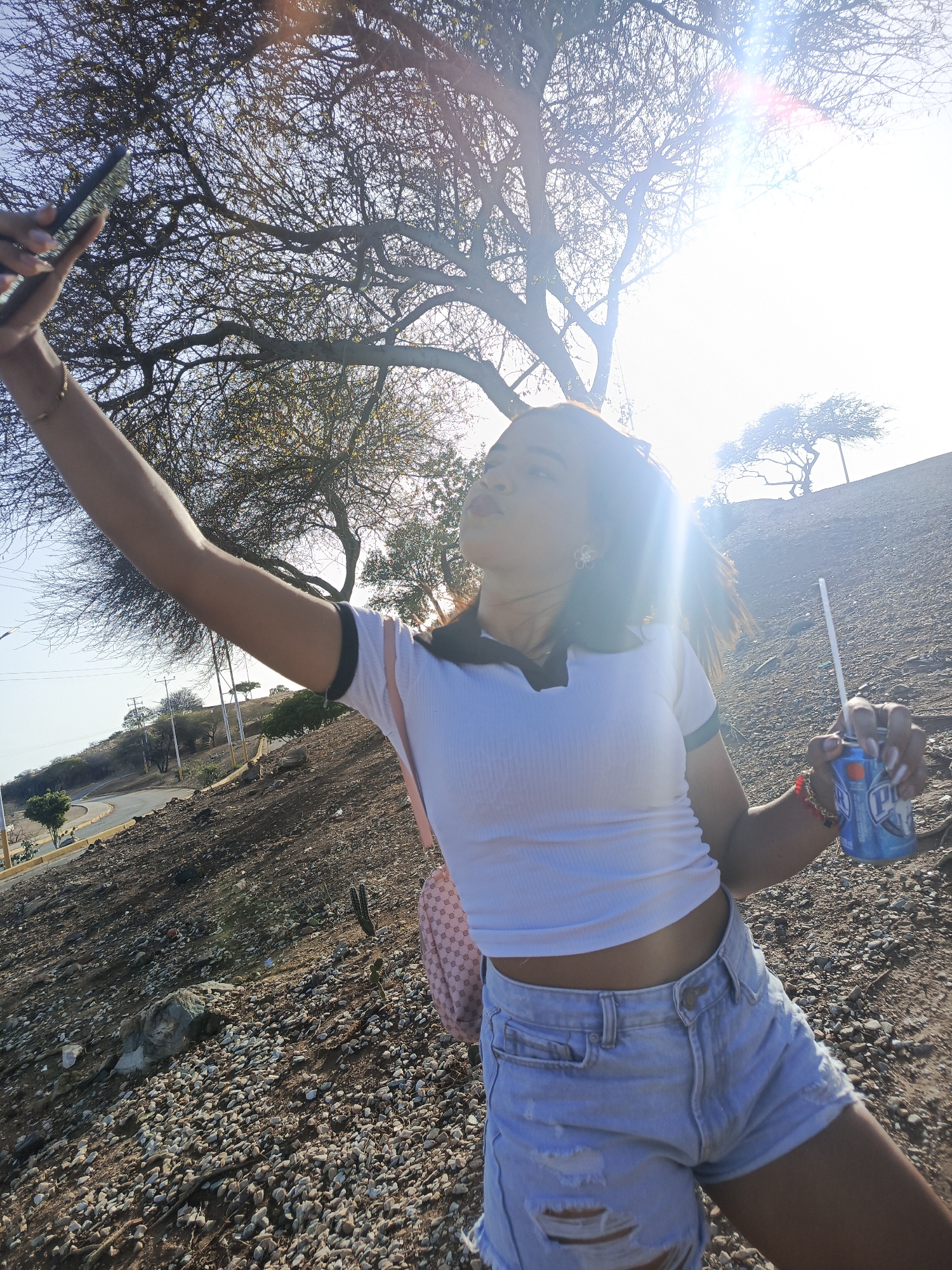 A young woman is captured taking a selfie outdoors in a bright, arid environment, likely Juan Griego, Venezuela. She is shown from the chest down, extending her left arm to hold a dark smartphone and capture the image. In her right hand, she holds a blue drink can with a white straw, prominently displaying the letter "P" on its label.
She is wearing a white ribbed crop top with a black-trimmed collar and light blue ripped denim shorts. A light-colored backpack is visible on her right shoulder. Her expression, partially obscured by the strong sunlight and lens flare, appears relaxed as she looks towards the phone.
The background depicts a dry, hilly landscape covered in small stones and dirt. Sparse trees with intricate, mostly bare branches are scattered across the terrain, indicative of an arid climate or dry season. A winding asphalt road, marked with white and yellow lines, ascends a hill on the left side of the frame, with utility poles visible alongside it. The sun, positioned behind and to the upper right of the subject, creates a significant lens flare and casts harsh light, resulting in a bright, somewhat washed-out sky. The scene suggests a warm, clear day in a natural, semi-deserted setting.