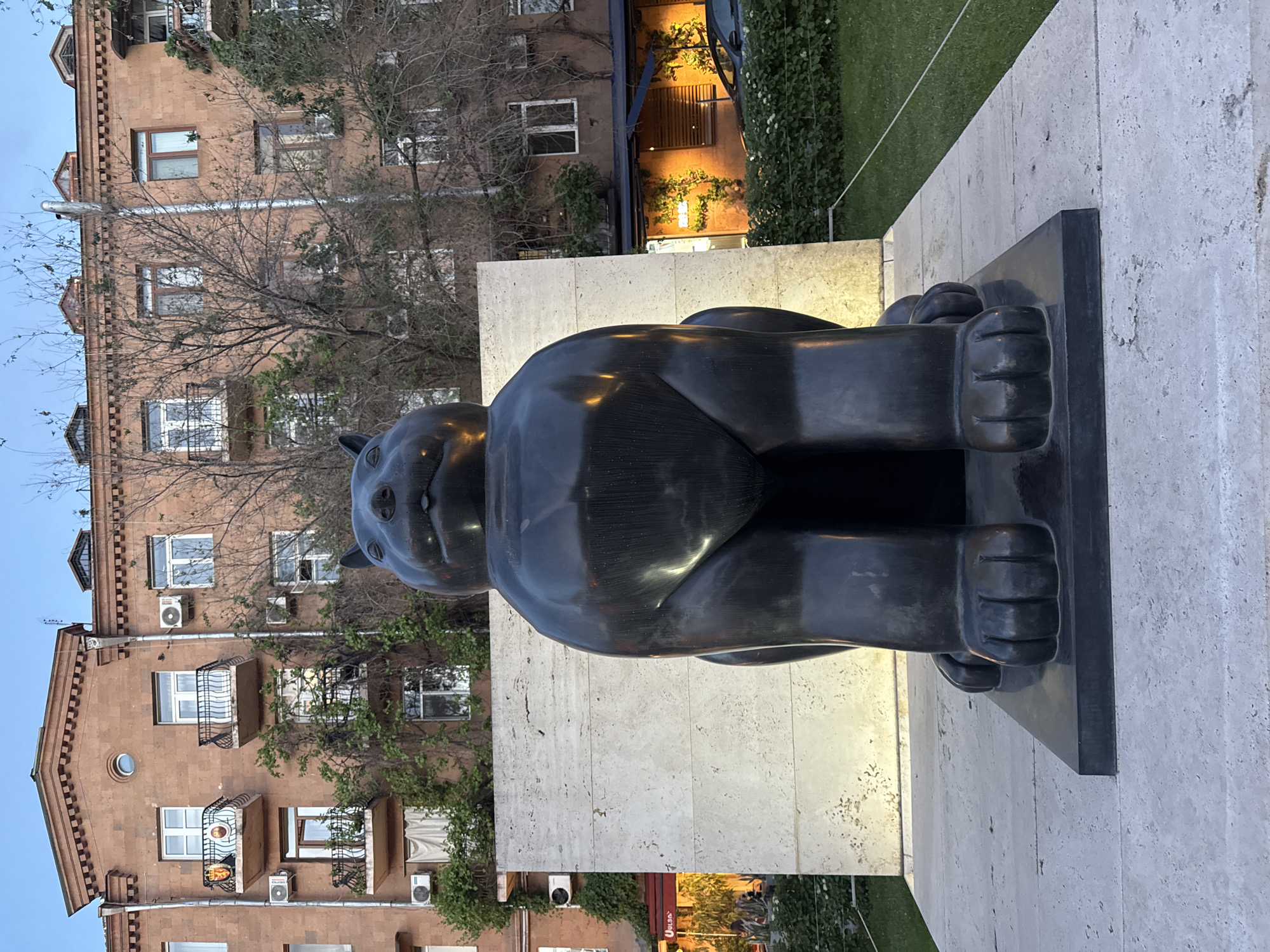 The image presents a large bronze statue of a cat, squatting on a black plinth that sits atop a beige-colored stone block. The sculpture is positioned outdoors in an urban environment, as evidenced by the background buildings. The backdrop features a multi-story brick building with windows, balconies, and air conditioning units, characteristic of residential or commercial structures. Some greenery is visible, including trees and vines, adding an organic element to the urban setting. The presence of the cat statue indicates a public space or a designated art installation area. Based on its distinctive design and the surrounding architecture, this is likely the Gata Sculpture in Yerevan, Armenia.