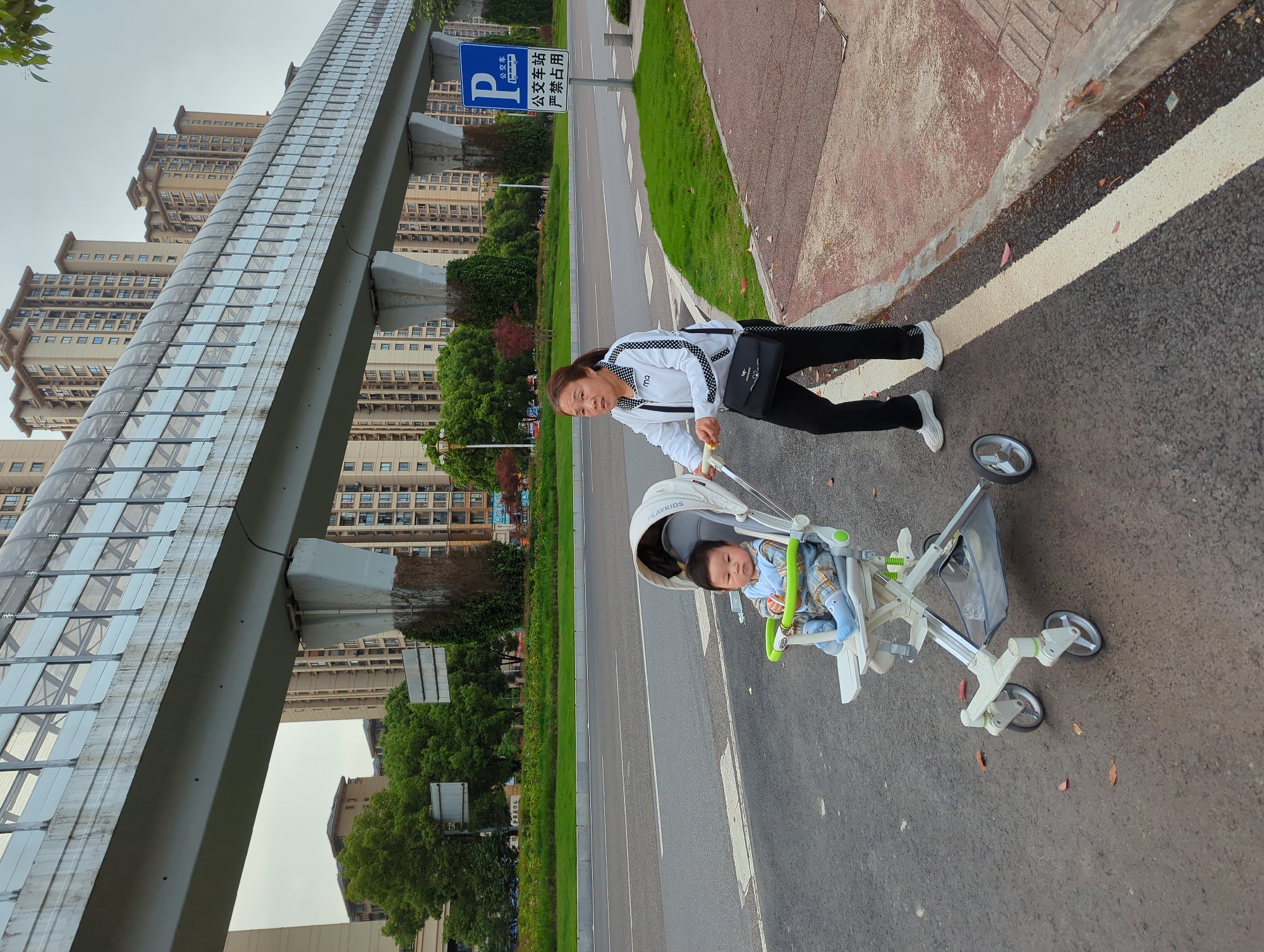 An outdoor, slightly rotated, full-shot photograph captures a woman pushing a child in a stroller on a sidewalk in an urban setting in Jiangjin, China. The woman, positioned on the right side of the frame, wears a white and black track jacket with "ma" written on it, black pants, and white sneakers, carrying a black shoulder bag. She holds the handle of a light grey and white "PLAYKIDS" brand stroller, in which a young child, dressed in a plaid shirt and light blue clothing, sits calmly, looking towards the right.
They are on a paved sidewalk, which transitions from a reddish-brown section to a darker asphalt path with a white dividing line. To their left, a wide, empty road extends into the distance, flanked by a strip of green grass and trees. In the background, numerous tall, multi-story residential buildings rise under an overcast sky. A prominent, modern-looking pedestrian overpass, constructed with glass and metal, spans across the upper left portion of the image, reflecting the sky and surrounding buildings.
Near the road, behind the woman, a blue rectangular road sign is visible. It features a white 'P' symbol, a bus icon, and Chinese characters that read "公交车" (bus), and below that "公交车站 严禁占用" (Bus stop, strictly forbidden to occupy). The overall scene depicts a quiet moment of a parent and child walking in a bustling city. The time of day appears to be daytime, with soft, diffused light suggesting an overcast or mildly cloudy condition.