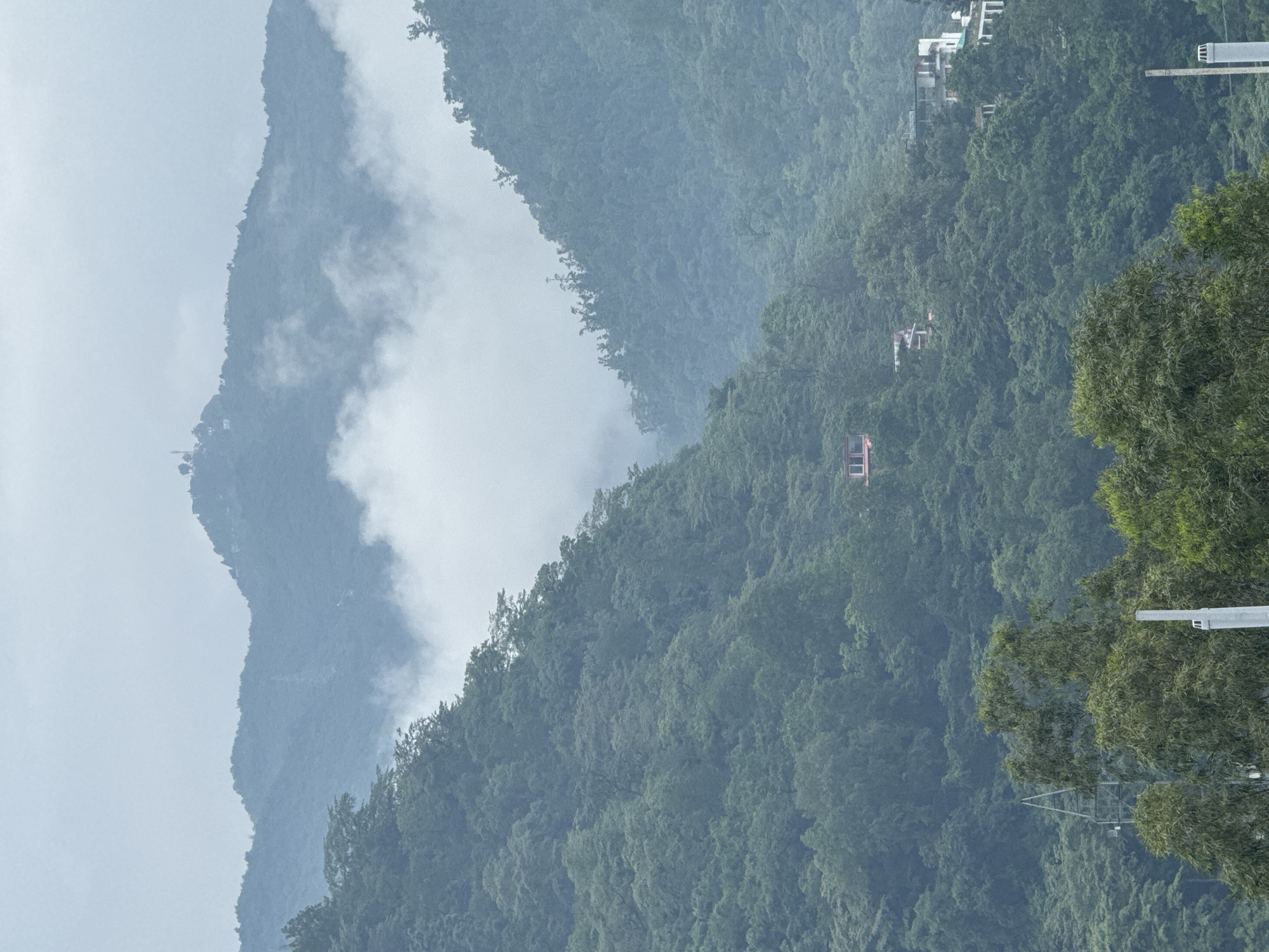 The image depicts a foggy, overcast day in Rishikesh, India, characterized by dense green hills covered in lush vegetation. In the mid-ground, several buildings are visible nestled into the hillsides. A prominent red-roofed structure, possibly a house or a small resort, is situated on the left side of the frame. Further to the right, other buildings with white facades and angular roofs are scattered amongst the trees. In the foreground, the tops of trees and some man-made structures, including what appears to be a utility pole or tower, are visible. The distant mountain peaks are partially obscured by soft, swirling clouds or fog, creating a serene and somewhat mystical atmosphere. There are no people or animals discernible in the image. The overall scene conveys a sense of a remote, natural setting within a developed area.