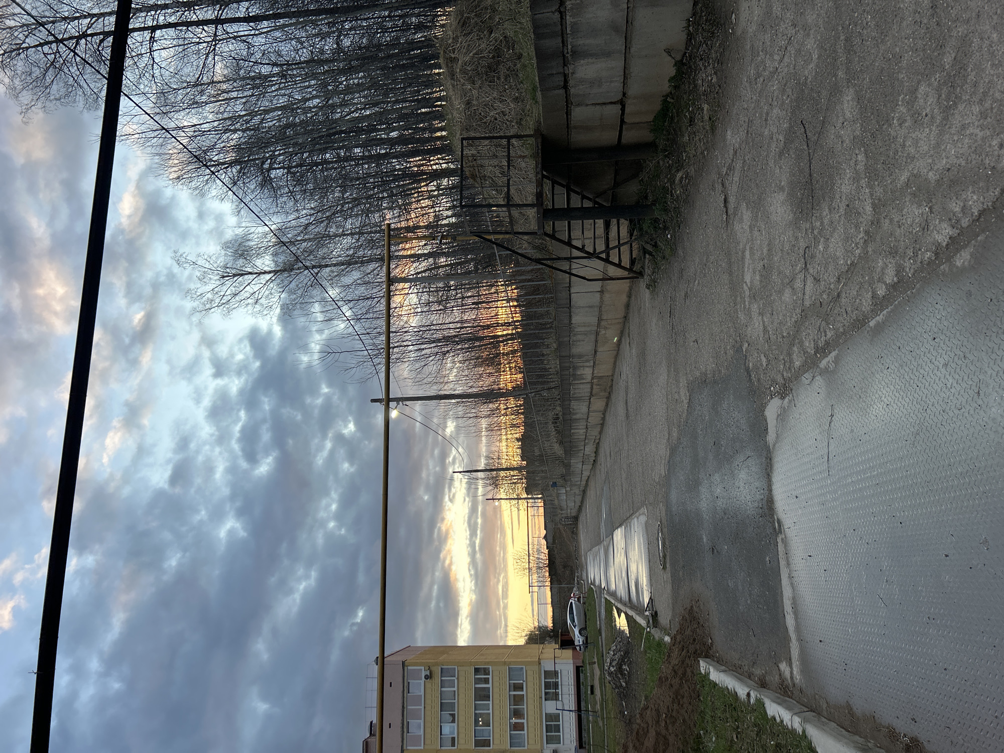 The image depicts an outdoor scene in Noviy, Russia, captured during either sunrise or sunset, evidenced by the vibrant colors on the horizon. The sky transitions from dark, heavy clouds in the upper portion to a brilliant blend of orange, yellow, and soft purple hues where the sun's light breaks through.
In the foreground, a paved path, possibly concrete or asphalt, stretches across the scene, appearing wet in areas, suggesting recent rain. A section of diamond-plate metal flooring is visible on the far right. Parallel to the path, a low wall constructed from concrete blocks runs along the right side, separating the path from a slightly lower, overgrown embankment. Beyond this, a dense line of bare trees, indicative of late autumn or winter, forms a stark silhouette against the brightly lit sky.
On the left side of the frame, a multi-story building with a light yellow facade and multiple rectangular windows is visible, likely a residential or administrative structure. A light-colored car is partially parked near this building. Several dark utility poles and overhead lines crisscross the sky, adding to the urban or semi-urban setting. A dark, metal, cage-like structure or set of stairs stands near the concrete wall and trees. No people are visible, and no specific activity is taking place. The scene conveys a quiet, somewhat utilitarian atmosphere, punctuated by the striking natural beauty of the sky. No visible text is present.