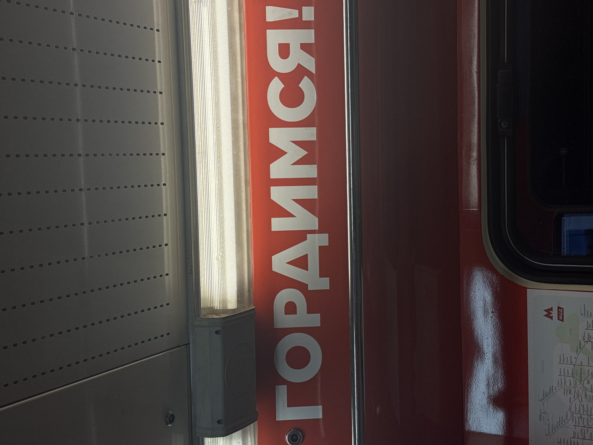 The image depicts the interior of a Moscow Metro train car in Russia. A portion of the side wall is visible, constructed from perforated grey metal panels. Attached to the wall is a fluorescent light fixture. Adjacent to the light fixture, a red panel displays the text "ГОPДИMCЯ!" (Gordimsya!), meaning "We are proud!" A portion of the train car door and window can be seen on the right, next to a map of the Moscow Metro system.