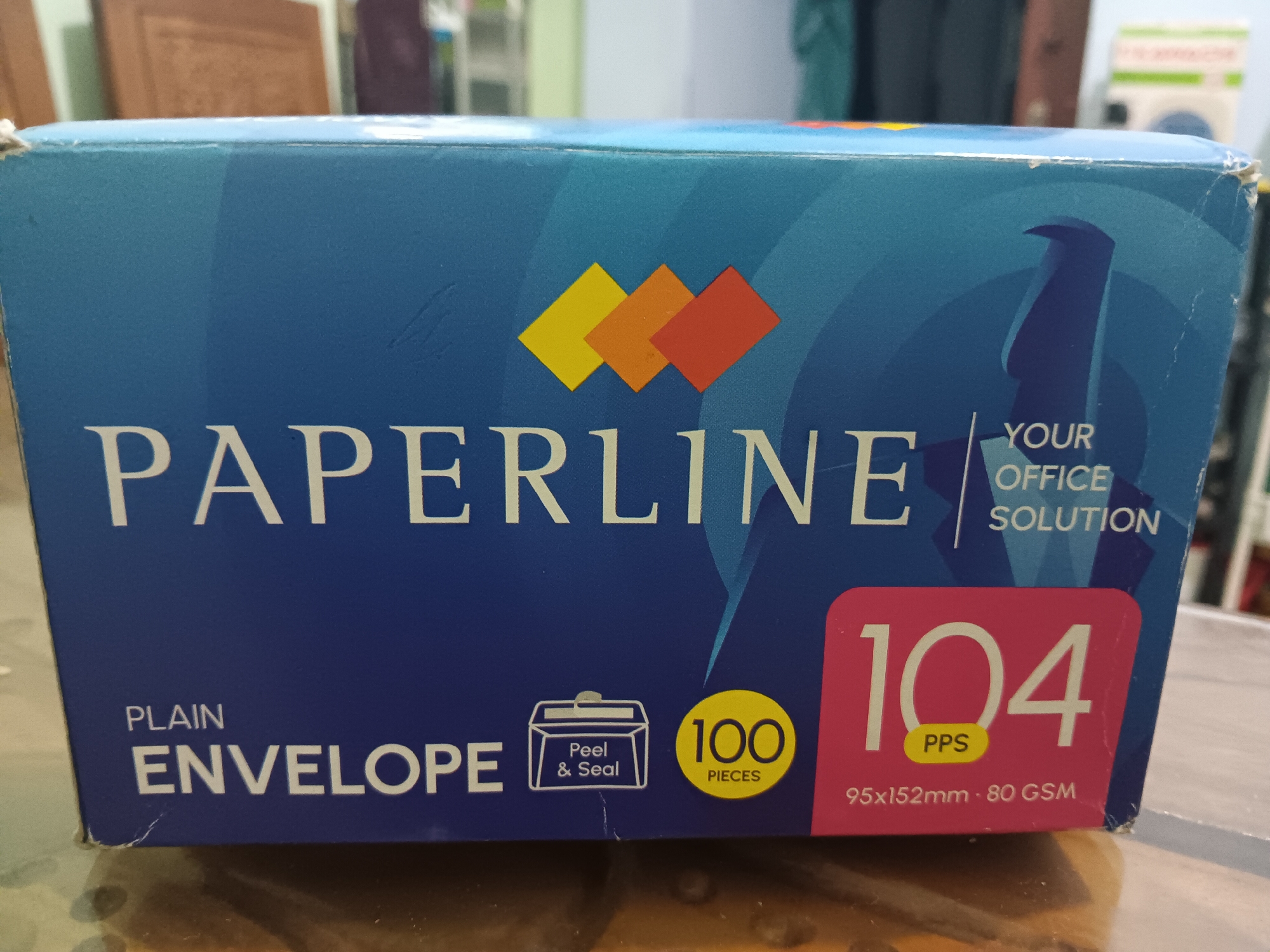 This is a photograph of a box of Paperline brand plain envelopes. The box is predominantly blue with white lettering and orange, yellow, and red square graphics. The front of the box displays the brand name "PAPERLINE" in large white letters. Below that, it reads "PLAIN ENVELOPE" and shows a graphic of an envelope with "Peel & Seal" written inside. To the right of this graphic, there is a yellow circle with "100 PIECES" written in black. Further to the right, a large pink rectangle displays "104 PPS" in white lettering and "95x152mm - 80 GSM" in smaller white lettering. The box appears to be sitting on a table with a wooden surface. The background is blurred but suggests an indoor setting with wooden furniture and shelves. The image does not contain any information to determine its location in Batursari, Indonesia.