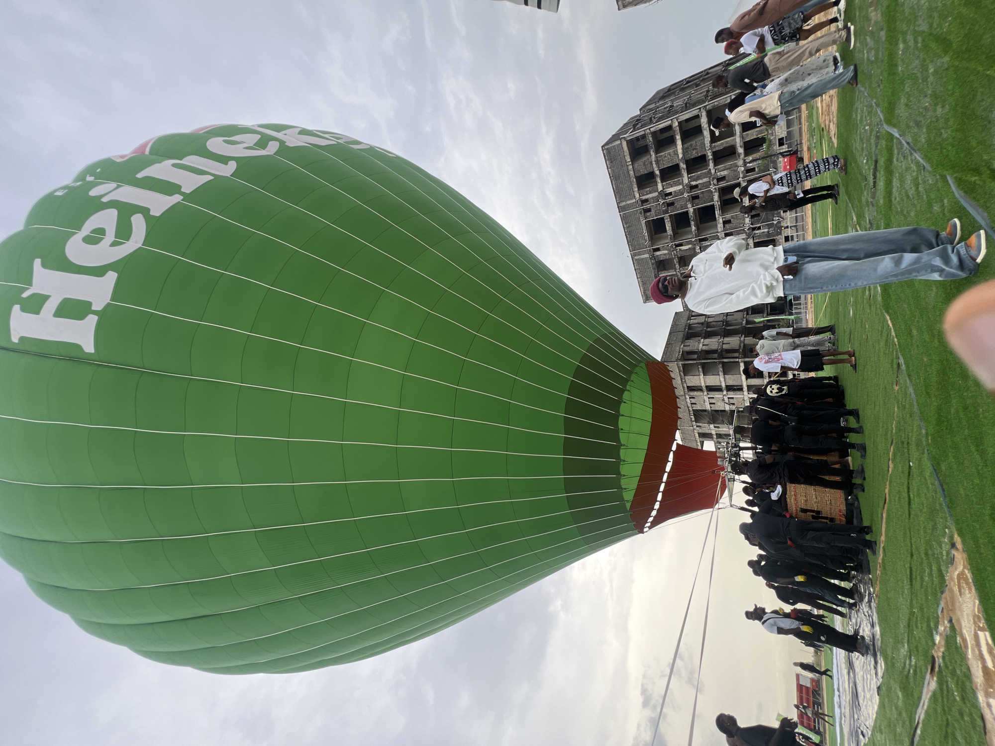 A large green hot air balloon with the word "Heineken" printed on it is being prepared for takeoff on a grassy field. A group of people, primarily dressed in casual attire, stand around the balloon. Some appear to be crew members, while others seem to be spectators. In the background is an unfinished or abandoned high-rise building, which contrasts with the vibrancy of the balloon and the grassy field. The scene is taking place in Lagos, Nigeria. The sky is overcast, indicating it is either early morning or late afternoon.