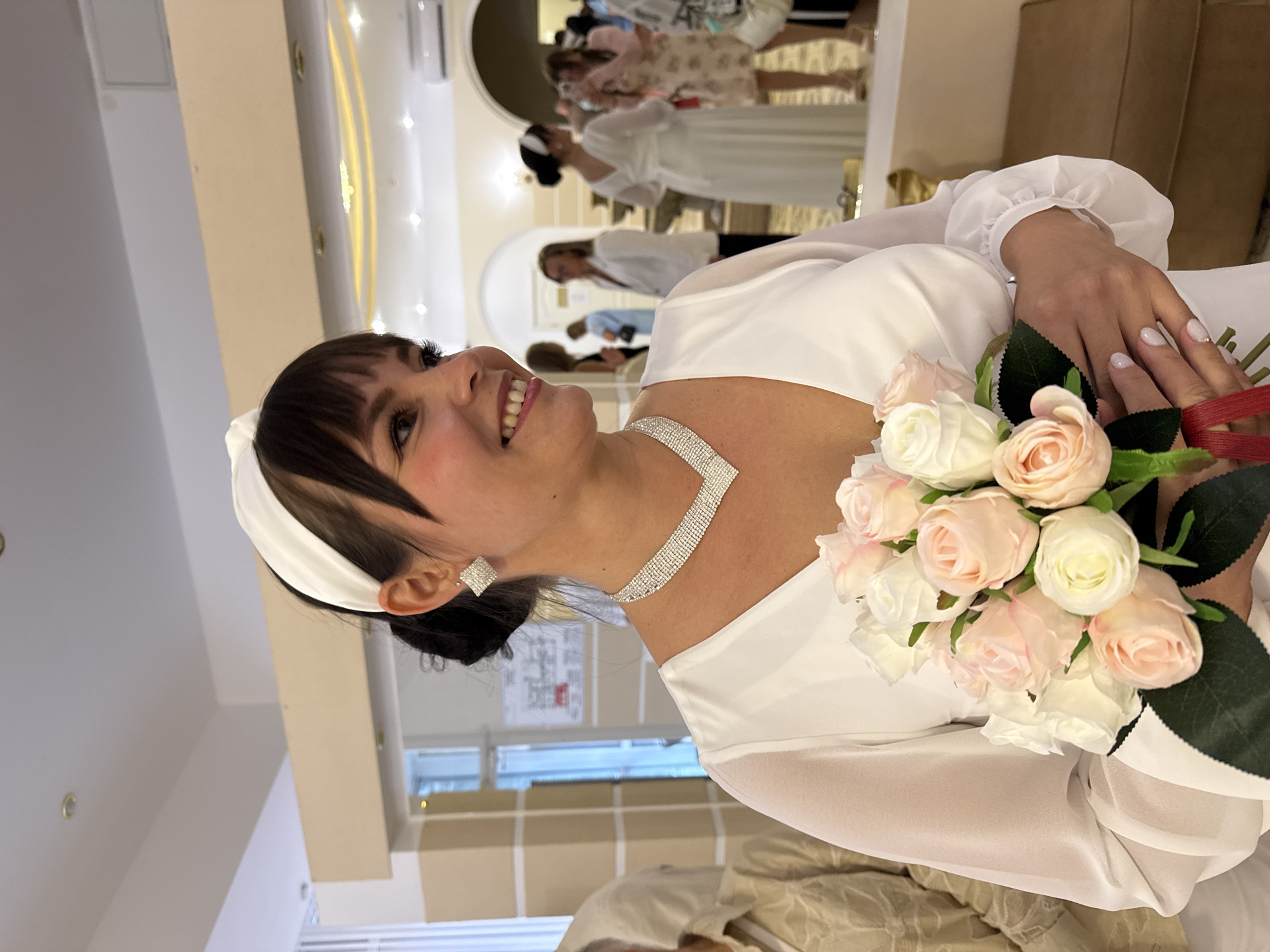 A woman with dark hair, wearing a white headband, white dress, and diamond jewelry, holds a bouquet of pink and white roses. She is smiling and looking to her right. The background shows a room with mirrors, lights, and other people, suggesting an indoor event such as a wedding reception.