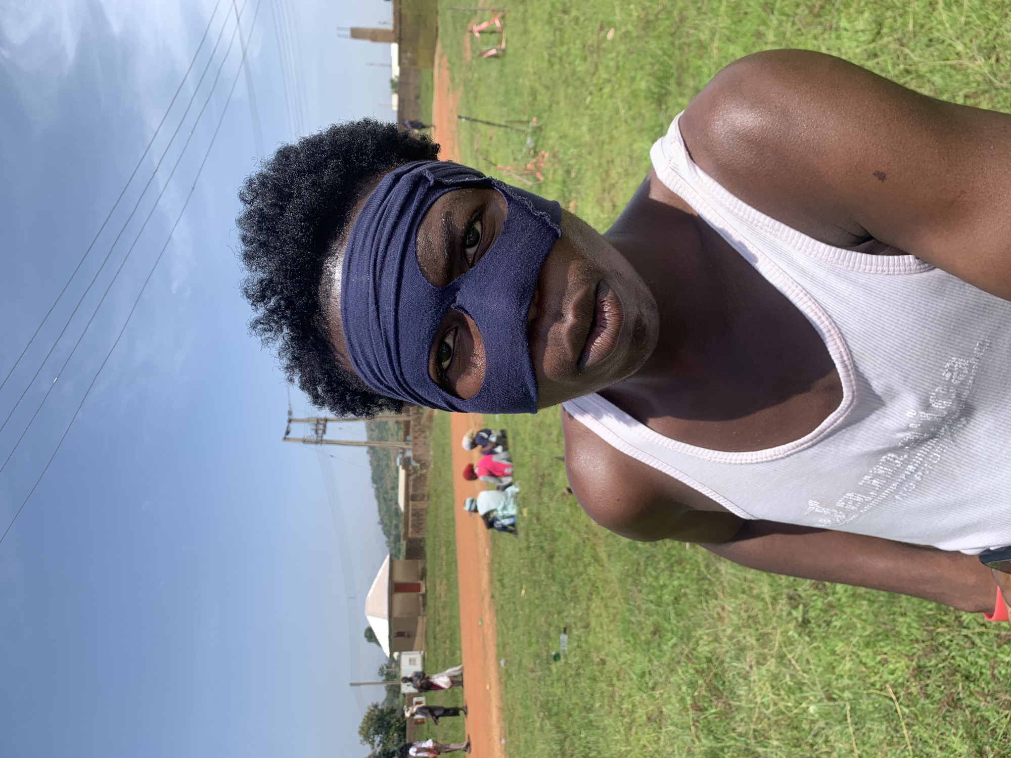 A medium shot shows a person wearing a dark blue cloth mask that covers their forehead and the upper part of their face, with holes for the eyes. The person's short, dark, curly hair is visible above the mask. They are wearing a white tank top. The person is looking directly at the camera. The background is an outdoor scene with green grass. Several people are sitting in the background on the grass. To the left, there is a dirt path and a few buildings. Power lines are visible in the sky. The scene is likely in Gwafan, Nigeria.