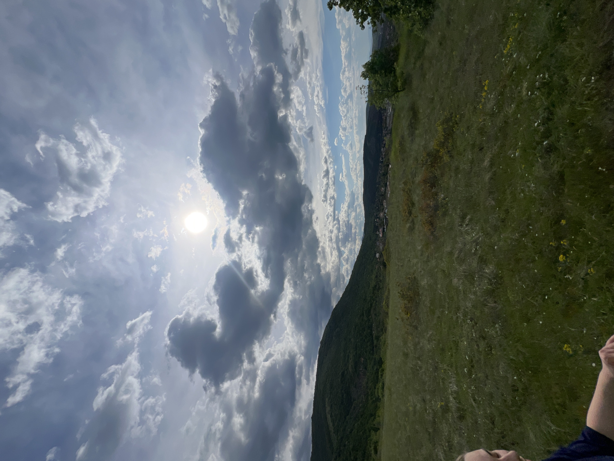 The image shows a landscape with a cloudy sky and a field. The sun is visible through the clouds on the left side of the image. A mountain range runs diagonally across the center of the image, separating the sky from a green field with scattered yellow and white flowers. On the side of the mountain in the distance are some buildings and trees. An arm and part of a face is visible in the bottom right corner, suggesting the photo was taken by a person. Based on the location information provided, this scene may be in the vicinity of Markovo, Bulgaria.