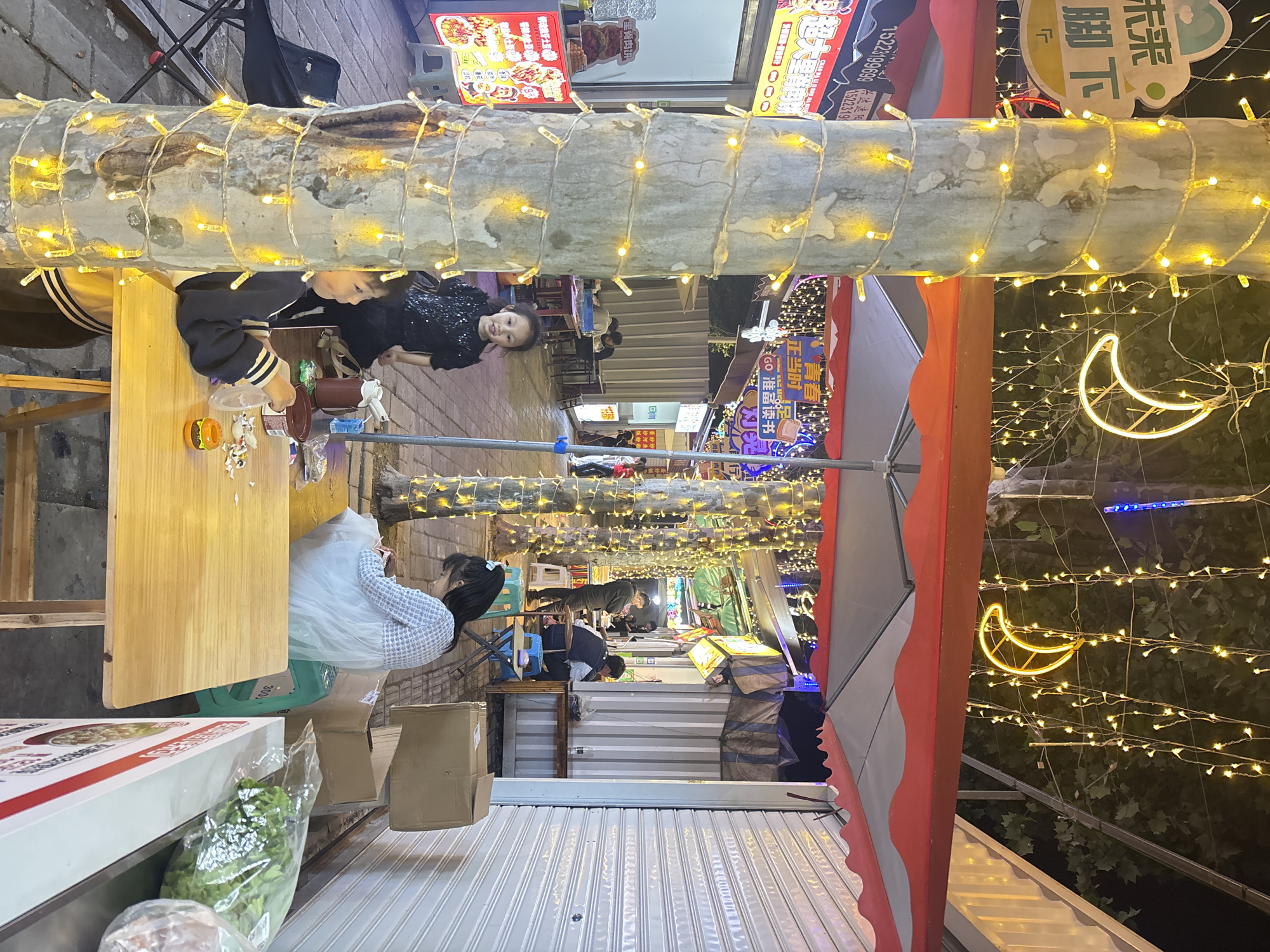 This nighttime image shows a street scene, likely in Jiangjin, China, based on the Chinese characters on the signs. A string of lights runs through the center of the frame, with people dining at tables on the left. Two women are visible, one in a black dress and another in a light-colored top. Food and drink are on the table. The street is lined with shops and stalls, lit with signs and displays. There are decorations including lights strung up along the street, and a crescent moon shaped light. There are several boxes and packages in the foreground, along with produce. Some of the signs contain phone numbers and descriptions of food items.