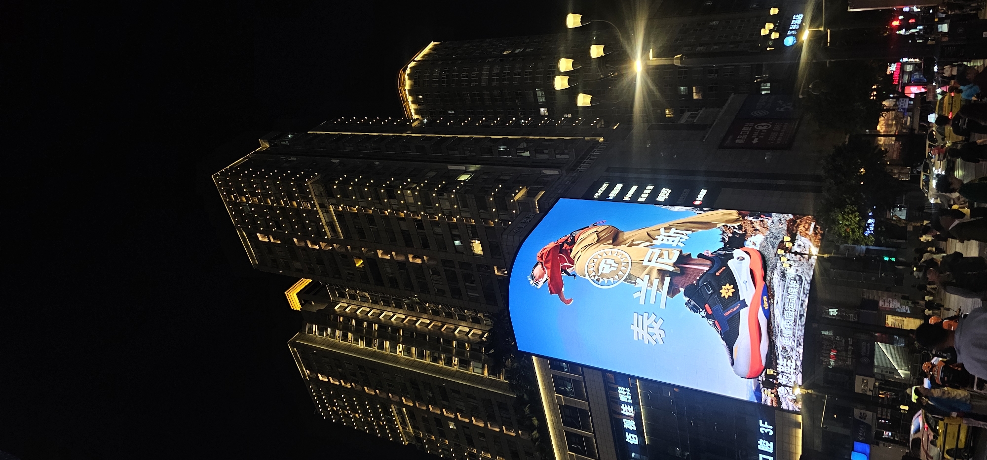 A nighttime view in the Yuzhong District, China, depicts illuminated high-rise buildings adjacent to a large digital display screen. The screen shows an advertisement featuring a person and footwear. Pedestrians and vehicular traffic populate the street below. Lighting from street lamps and building illumination is prominent.