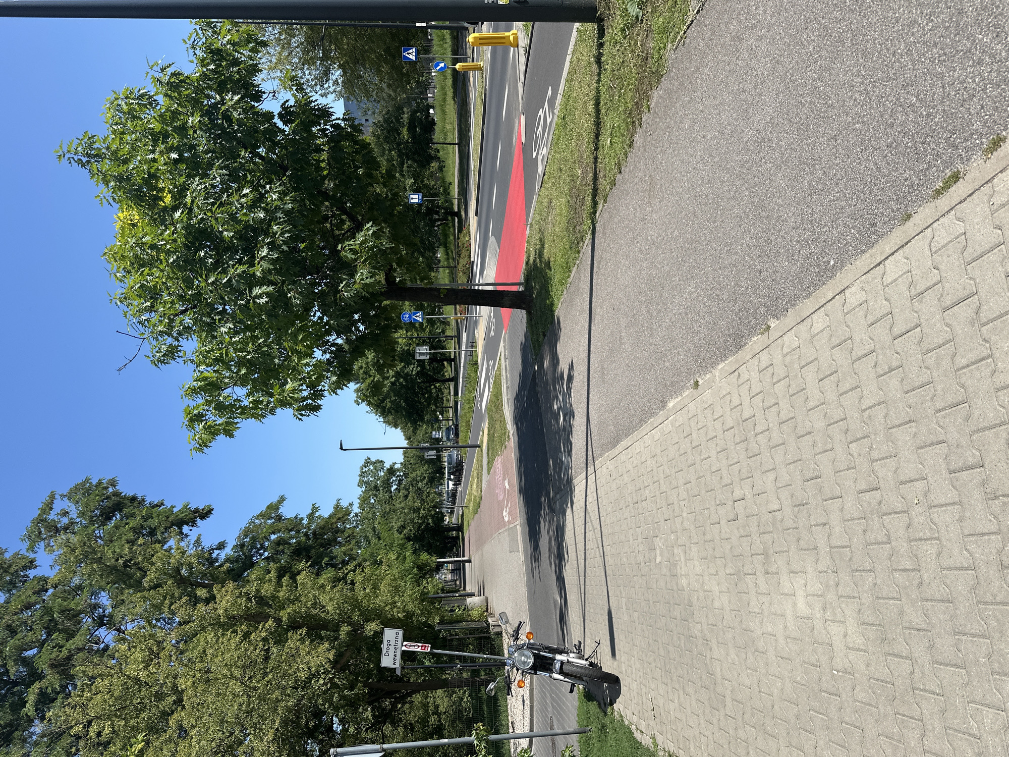 This image depicts a scene along a city street in Warsaw, Poland, on a clear, sunny day. A paved sidewalk, constructed with interlocking concrete pavers, runs along the left side of the frame, leading into the distance. A motorcycle is parked on the grass verge adjacent to the sidewalk, its kickstand supporting it.
To the right of the sidewalk, there is a road with designated lanes. A red painted area on the road surface indicates a specific zone, possibly for cyclists or a transition area. Road markings, including bicycle symbols, are visible on the asphalt. Several street signs are present: one indicating "Droga wewnetrzna" (internal road) with a prohibitory symbol, and others with blue and white traffic symbols, including a directional arrow and a pedestrian crossing sign. Yellow bollards are positioned along the roadside.
Lush green trees line the street, providing shade. In the background, vehicles are driving on the road, and buildings are visible in the distance. The overall environment suggests an urban setting with dedicated infrastructure for both vehicular and non-vehicular traffic.