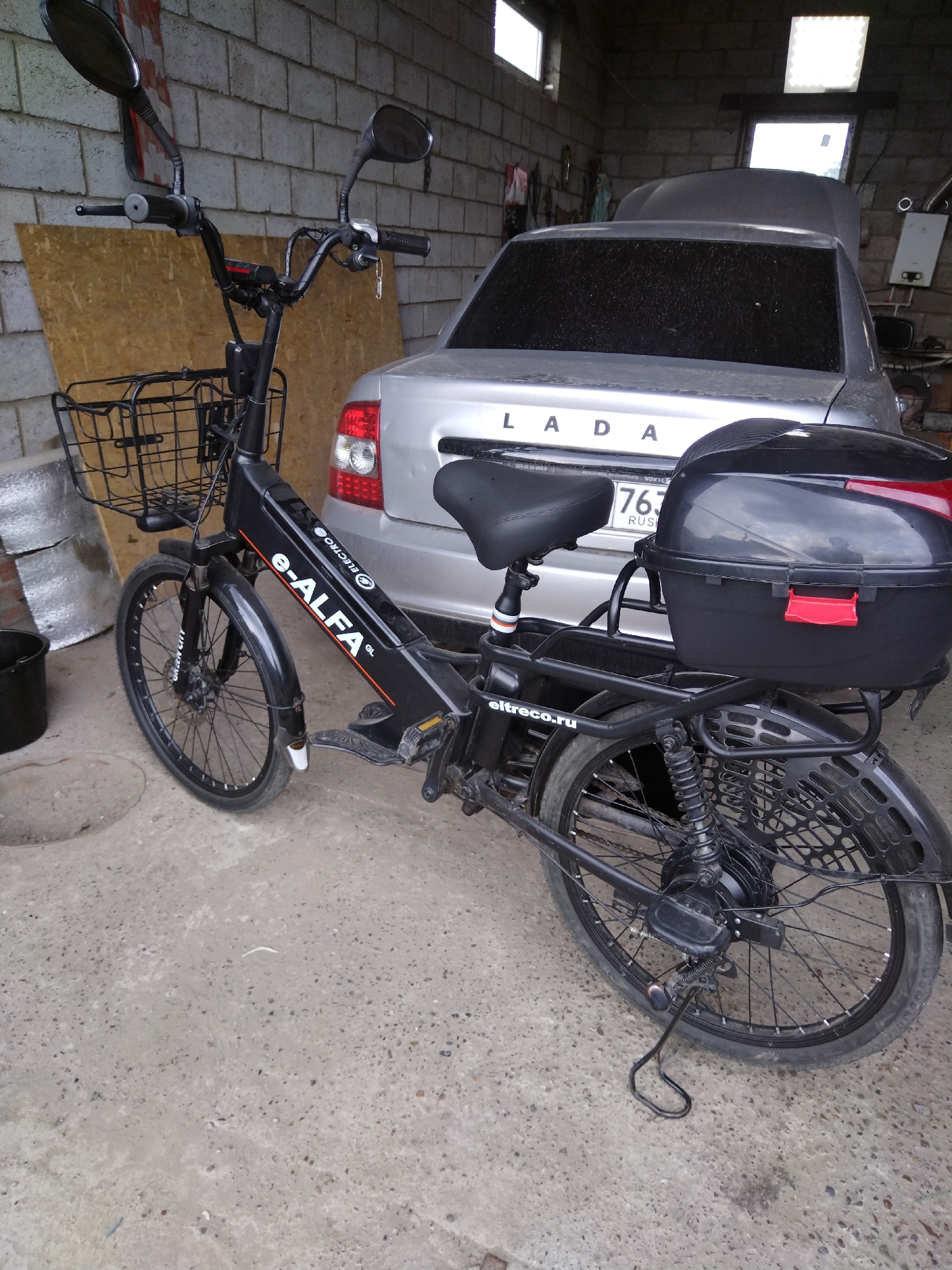 An electric bicycle, identified by the "e-ALFA" branding, is parked on a concrete floor indoors, possibly in a garage or workshop in Bazarnyye Mataki, Russia. The bicycle is black and features a front basket, a rear storage box, and dual mirrors. Behind the bicycle, a silver Lada car is partially visible, with its hood open. The environment is characterized by a brick wall, a wooden panel, and various indistinct objects in the background, suggesting an unfinished or utilitarian space. The lighting appears to be artificial, with a bright panel visible on the ceiling.