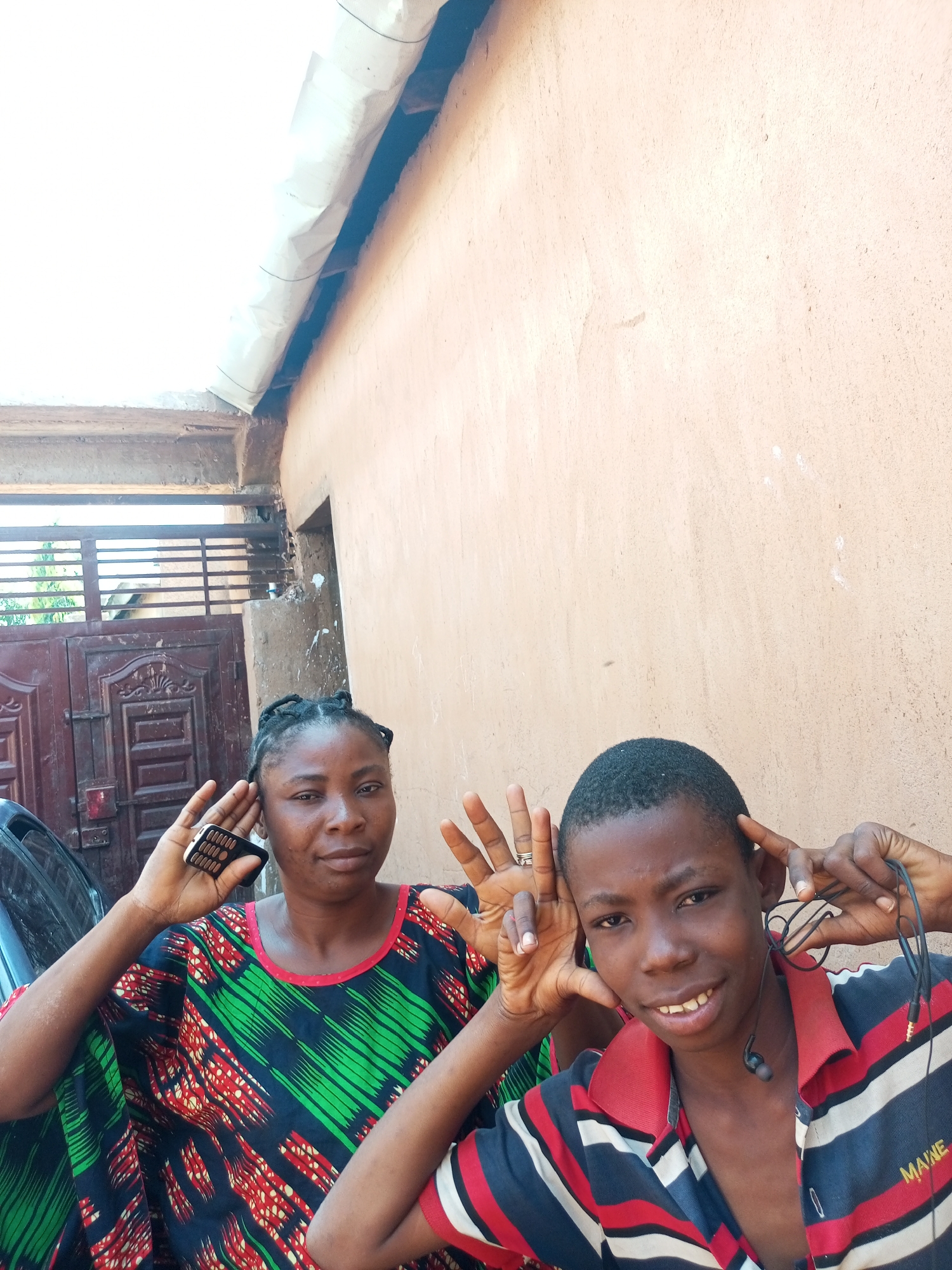 In Tempure, Nigeria, two individuals, an adult woman and a youth, are captured in a close-up portrait. The woman, on the left, wears a colorful, patterned kaftan and holds a mobile phone to her ear. She has braided hair and her fingers are posed near her temple. The youth, on the right, sports a striped polo shirt with headphones around his neck and makes a gesture with his fingers near his temples. Both individuals are looking towards the camera. They stand in front of a textured, light-colored wall, with a dark metal gate and a portion of a building visible in the background. The lighting suggests an outdoor setting, possibly during the daytime.