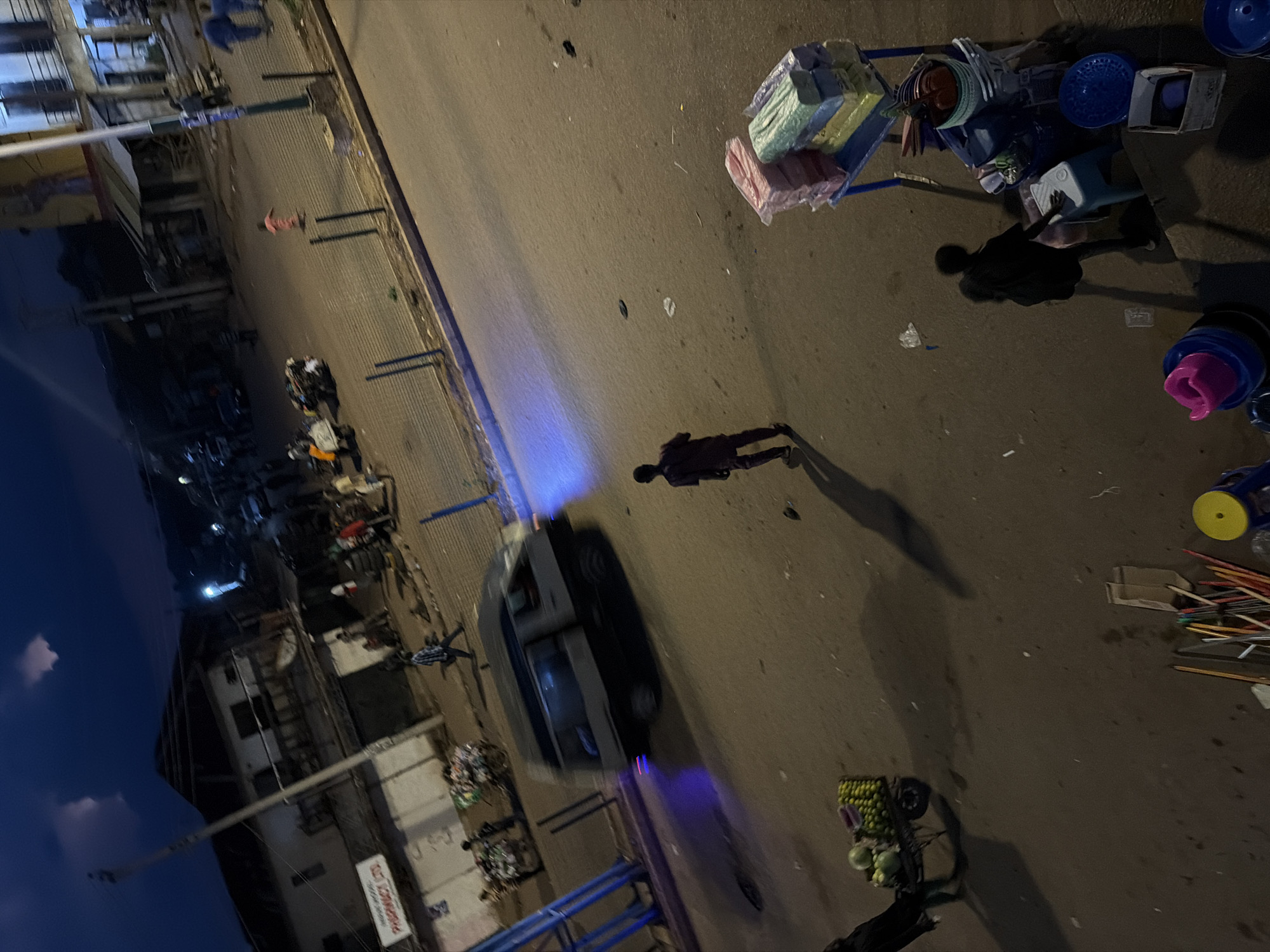 The image depicts a street scene at dusk in Bukuru, Nigeria. The sky is dark blue with wispy clouds, indicating late evening. A street is paved and appears somewhat worn, with scattered debris.
In the foreground, a person is seen from behind, wearing dark clothing and walking away from the viewer. To the right, a stand displays stacks of colorful items, possibly packaged goods or textiles, in pink, green, and yellow. Below this, various plastic containers and household items are visible, including blue and red bowls, and a blue plate. A small stool sits near the stand, with another person in dark clothing partially visible sitting on it, wearing a backpack. A shopping cart laden with green fruits, possibly melons and limes, is to the left, being pushed by a figure also wearing dark clothing.
Further into the scene, a dark-colored van is driving down the street, its tires blurred by motion. A blue light emanates from the rear of the van, casting a glow on the road. A figure in a reddish outfit is walking across the street in front of the van. On the left side of the street, a building with a sign that reads "PHARMACY LTD." is visible. Several people are gathered near this building, and a cart with stacked goods is parked nearby. In the background, other buildings line the street, with some streetlights and the glow of shop windows illuminating the area. Several figures are scattered throughout the midground and background, some walking and others standing. A few vehicles are also visible in the distance.