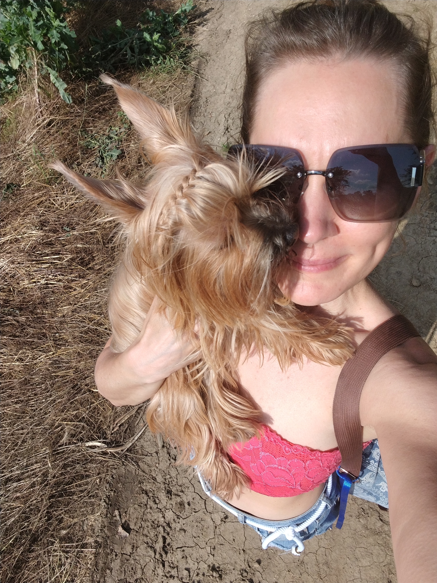 A woman is holding a Yorkshire Terrier while standing outdoors. She is wearing sunglasses, a pink lace bralette, and denim shorts. The ground appears to be dry and cracked, with some dry vegetation and green plants in the background. There is no indication of the location in Fyodorovskaya, Russia in this image.