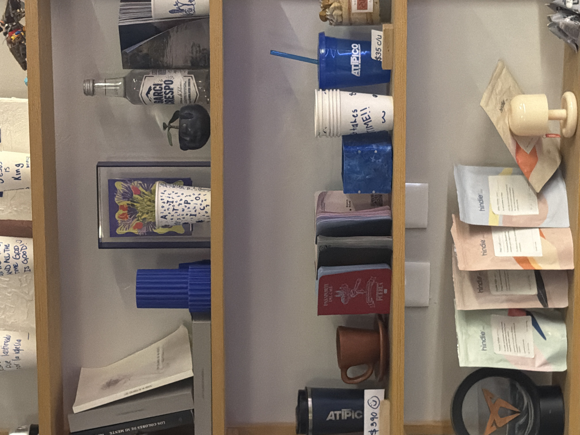 The image shows a wooden shelf filled with various items. On the left shelf, there is a bottle labeled "GARCI RESPO", books titled "LOS COLORES DE MI MENTE", and papers with handwritten notes. The right shelf contains a blue "ATIPICO" cup priced at "$35 c/u", coffee cups, a red "PASAPORTE" (passport) of "PUEBLA", packages labeled "hindie", a brown mug, and a black "ATIPICO" thermos priced at "$390". The overall environment is indoor, suggesting a shop or home in San Bernardino Tlaxcalancingo, Mexico.