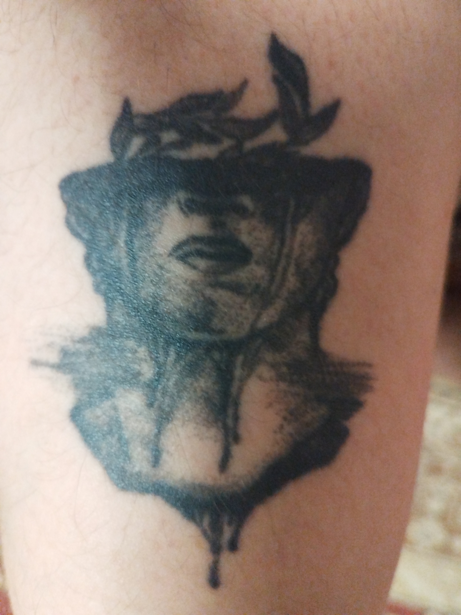 A tattoo of a classical bust wearing a laurel wreath is visible on a person's arm. The bust appears to be crying or melting, with black ink dripping down from its face. The tattoo is rendered in black and grey tones, with areas of heavy shading and detail. The background of the image is out of focus, showing skin and a textured surface, possibly fabric. The tattoo is the central focus of the image.