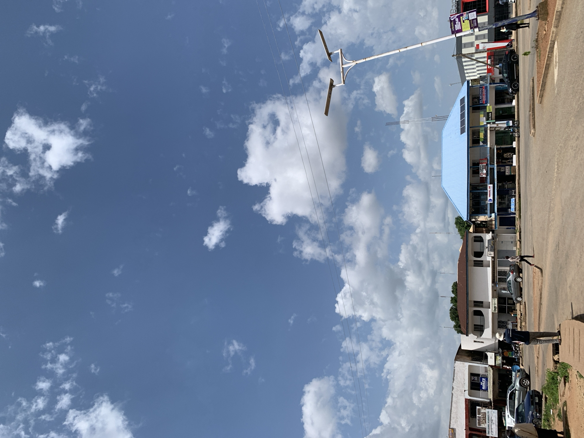 The image depicts a street scene in Jos, Nigeria, under a bright blue sky dotted with white, cumulus clouds. Several buildings line the street, displaying a mix of commercial storefronts. On the left, a white building has signage including "DIRAST UNIQUE LURIA" and "EMOY GLOBAL RESOURCES LTD." Next to it is another white building with a dark awning and signs for "MANLIS" and "COMMERCIAL BANK." Further down the street, a prominent building features a light blue, corrugated metal roof and multiple windows. Signs on this building include "m-bile" and "ST. ANTHONY SHOW ROOM." A black car is parked in front of this building. On the right side of the image, another building displays a red and black facade with "Bank" visible on a sign.
Several people are visible in the scene. In the foreground, a person wearing a light-colored hat and dark clothing stands on a raised concrete platform, facing away from the camera. To their left, a dark blue car is parked. Further down the street, a person in a dark shirt and light pants walks across the road. Another individual is seen walking from right to left in front of the blue-roofed building. A motorcycle is also present near the blue-roofed building. In the distance, a modern street lamp with two solar panels on top is visible. Overhead, thin electrical wires stretch across the frame. The road surface appears to be paved, with some areas of dirt and gravel along the edges. The overall atmosphere suggests a bustling urban environment on a clear day.