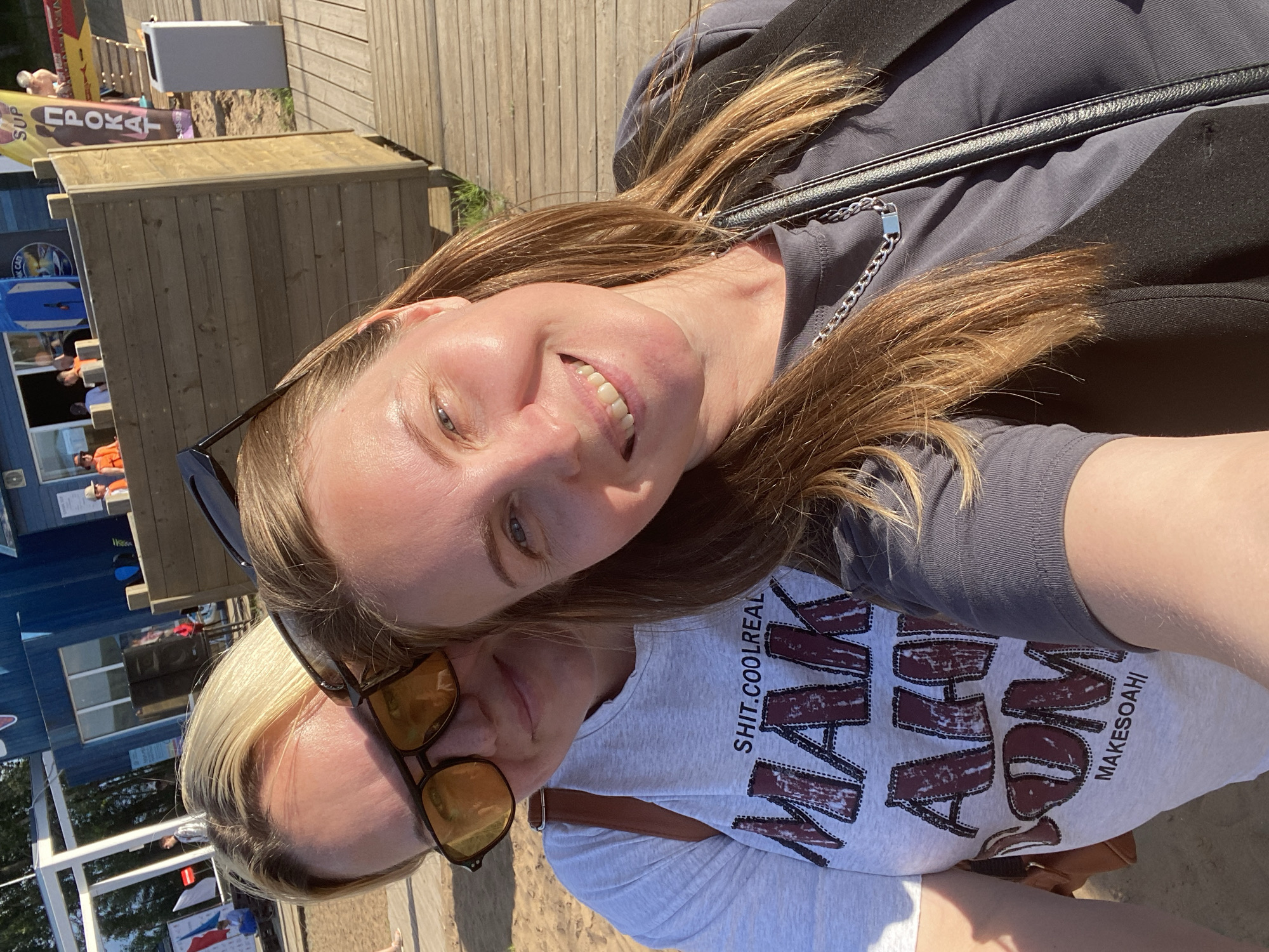 Two women are taking a selfie outdoors on a sunny day in Chaikovsky, Russia. The woman in the foreground is on the right, smiling at the camera. She has long, light brown hair and is wearing a grey t-shirt and a black vest. A silver chain necklace with a rectangular pendant is visible around her neck. The woman in the background is on the left, wearing sunglasses and a light grey t-shirt with a graphic print. She is also looking towards the camera and appears to be smiling. The background shows a beach area with sand, wooden structures, and a blue building. The overall scene suggests a casual outing, possibly at a recreational area or beach club.
