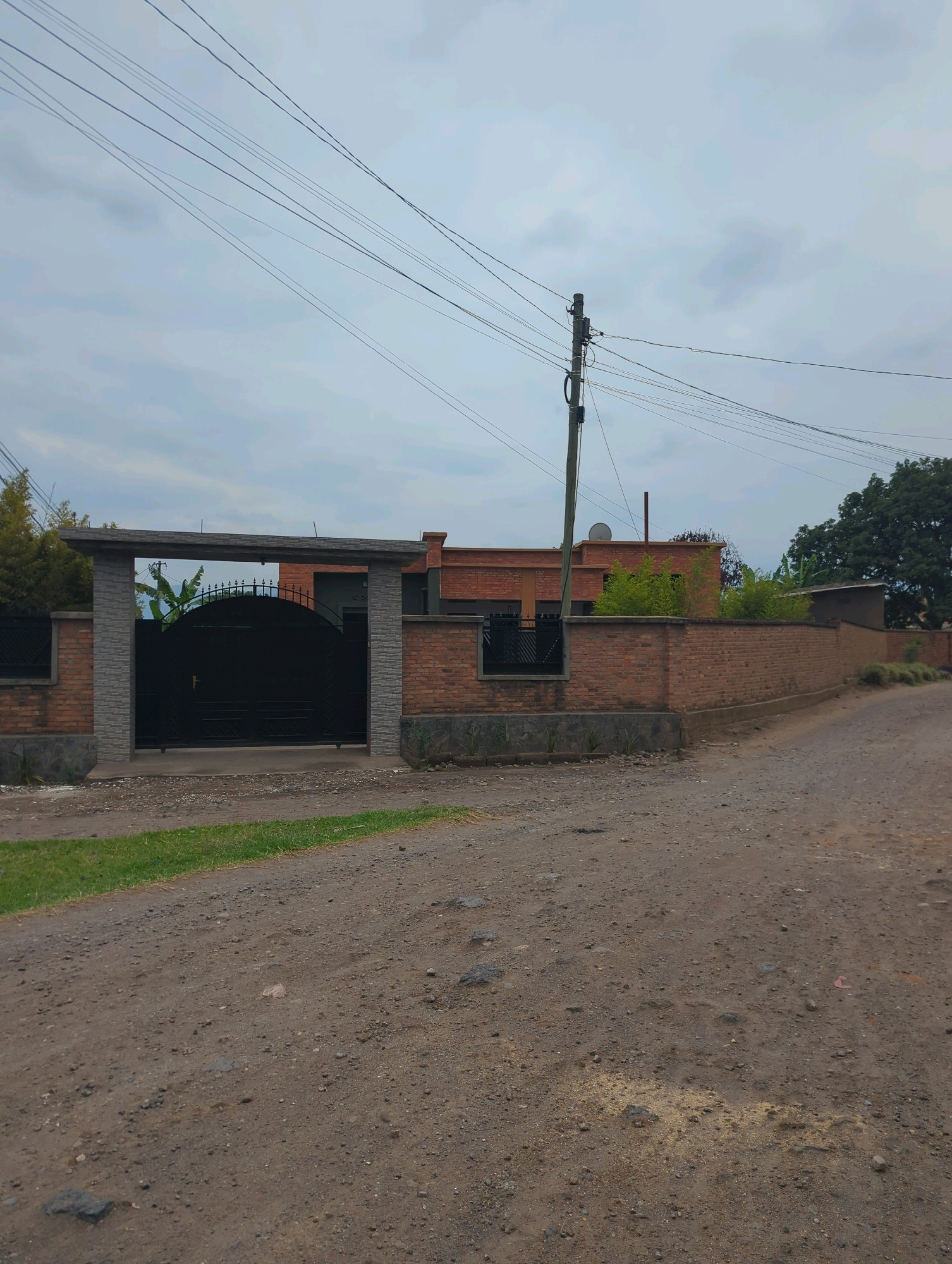 The image shows a street view in Musanze District, Rwanda, with a large brick wall and a black gate as the focal point. The gate is arched and ornate, with a gold handle. The wall is made of red brick and is several feet high, with a stone border at the base. Behind the wall, part of a brick building is visible, along with some trees and lush green vegetation. A wooden utility pole with numerous wires stretches across the sky, which is overcast. The foreground consists of a dirt road with some gravel and sparse green grass along the side. The overall impression is that of a residential property in a somewhat rural or suburban setting.
