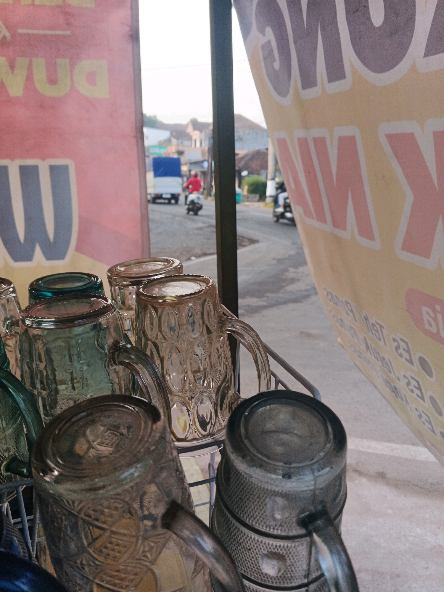 The image shows a rack of glass mugs, some transparent and others green, with decorative patterns, placed in front of a street scene in Semarang, Indonesia. In the background, a truck with a blue tarp is visible, and a person in a red shirt is riding a motorcycle. A large yellow banner with pink text, partially obscuring the view, is also present. The overall lighting suggests it is daytime.