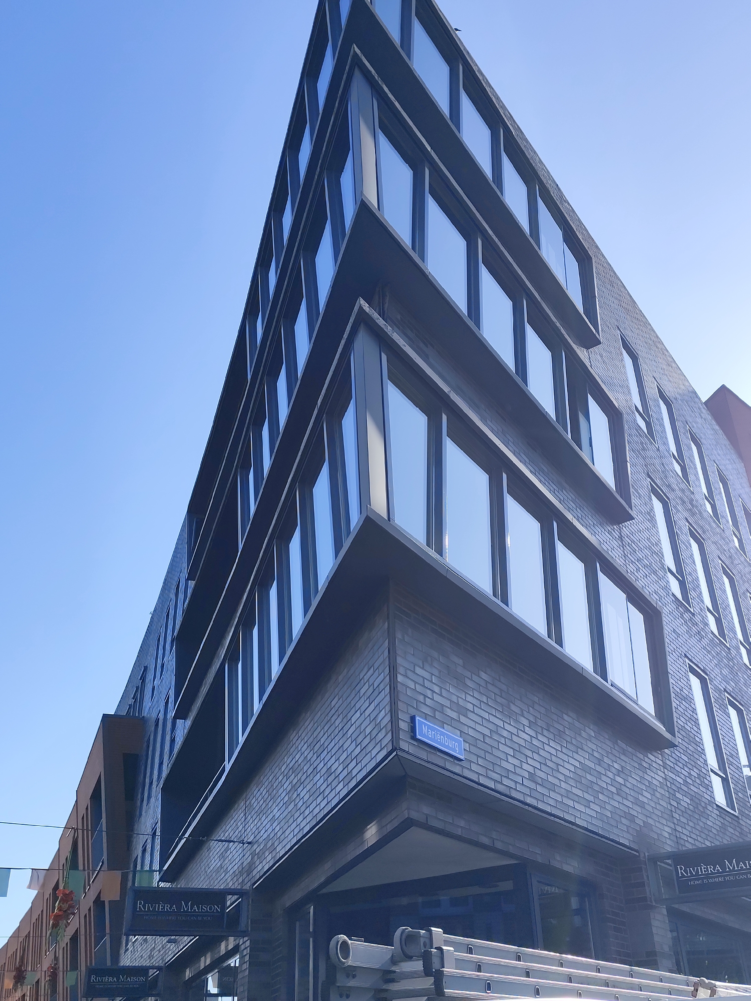 This is a low-angle, exterior shot of a modern building in Nijmegen, Netherlands, under a clear blue sky. The building features a facade of dark brick and large, reflective windows arranged in a grid pattern. A prominent corner of the building extends upwards and to the left, creating a sharp, angular silhouette. A blue street sign reading "Marienburg" is affixed to the brickwork near the corner. Below the sign, two black signs with white lettering that read "Riviera Maison" are hanging, one above the other, indicating businesses on the ground floor. One of the Riviera Maison signs has a tagline below the name. A section of a building with a brown facade and windows is visible to the left of the main subject building. In the foreground, part of a silver-colored ladder or scaffolding system is visible. The scene is devoid of people and the focus is on the architecture of the building and its immediate surroundings.