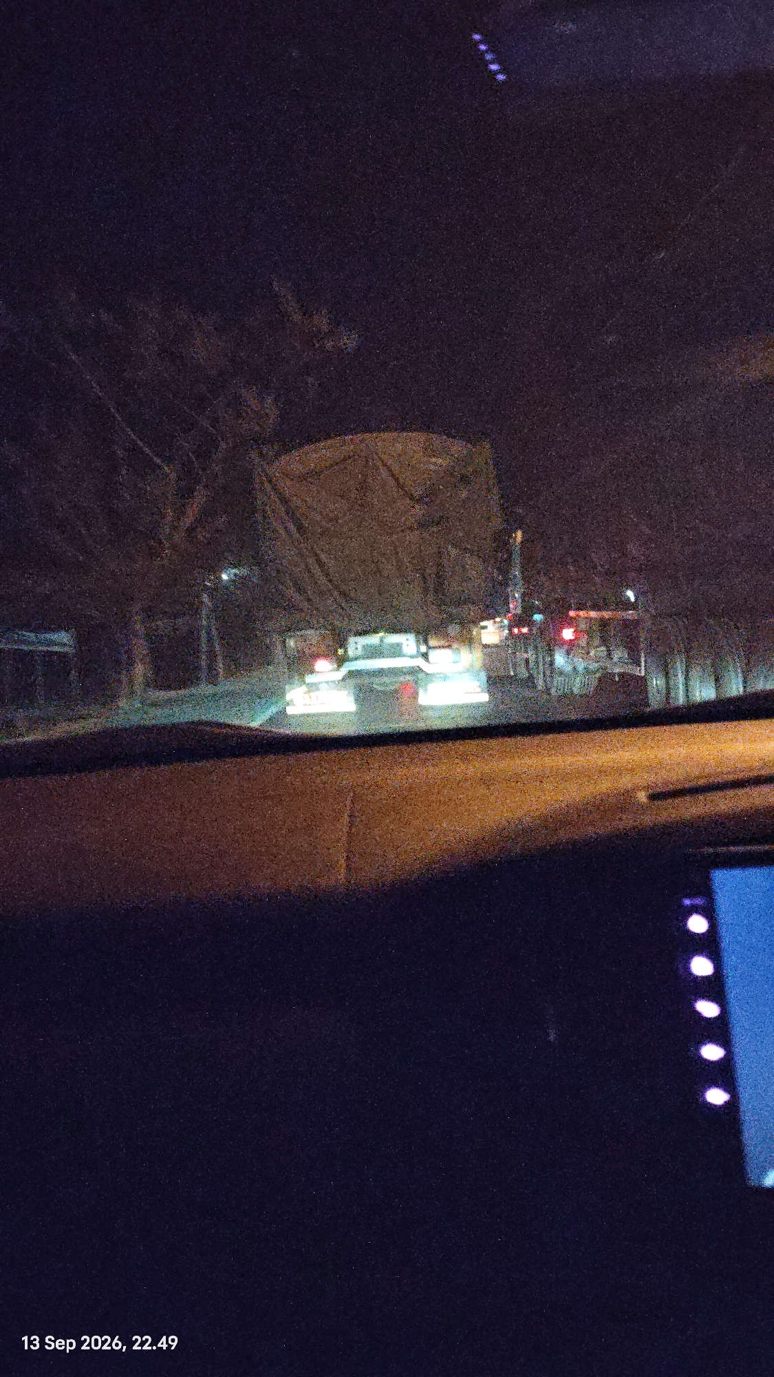 The image depicts a night-time scene from inside a vehicle looking out at traffic. The primary subject is a large truck covered with a tarp, its headlights illuminated. Several other vehicles with their lights on are visible behind it, suggesting a traffic jam or slow-moving traffic. The environment appears to be an urban or semi-urban area, with trees and structures visible in the background, all obscured by the darkness and glare from headlights. The image also includes a timestamp at the bottom left corner: "13 Sep 2026, 22.49". A portion of the vehicle's dashboard is visible in the foreground. There is no specific indication of the city of Pandanpancur, Indonesia.