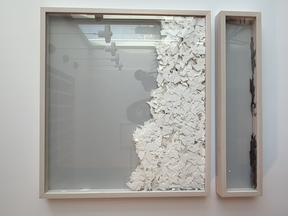 The image displays two contemporary shadow box art installations mounted vertically on a plain white wall within a modern interior of a high-rise building in Tel-Aviv, Israel.
The upper, larger rectangular shadow box features a light wood frame and contains a dense mass of small, irregularly shaped white ceramic or porcelain fragments, forming a jagged, wave-like landscape that fills approximately the lower two-thirds of the enclosure. The glass front of this frame exhibits reflections of the surrounding environment: two individuals are partially visible, one on the left appearing to hold a smartphone (likely the photographer), and another to the right, partially obscured, possibly seated at a desk. Multiple white, bell-shaped pendant lights are reflected, suspended from a ceiling, along with a large window on the far right revealing a distant cityscape and a segment of blue sky. A small amount of greenery (plant foliage) is also reflected near the bottom right corner.
Directly below the upper installation, a smaller, elongated horizontal shadow box, also with a matching light wood frame, is mounted. This lower box contains several dark, flat, irregularly shaped objects resembling small animal silhouettes, scattered across its base. These objects appear to be made of a dark, possibly metallic, material. The wall behind both art pieces is uniformly white.