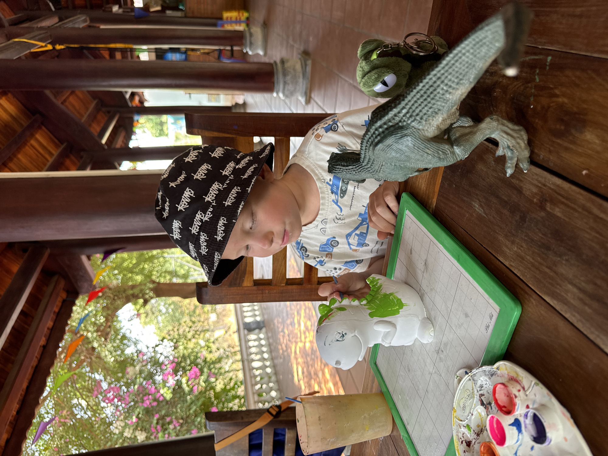 A young boy wearing a black patterned bucket hat and a white t-shirt with blue car designs is painting a small, white ceramic figure on a table in Mũi Né, Vietnam. He is using a paintbrush to apply green paint to the figure's head. Beside him on the wooden table is a green frog keychain and a grey toy dinosaur. A plastic palette with various colors of paint is also visible. The scene takes place outdoors, under a partially shaded structure with wooden beams and posts. Colorful decorations hang in the background, with glimpses of trees and flowers. A grid-lined board with a green border is placed under the ceramic figure, labeled "Phi Mã".