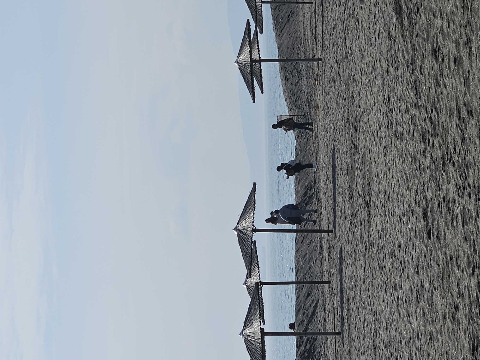 The image depicts a beach in Burgas, Bulgaria, characterized by dark sand. Several thatched umbrellas are lined up along the beach. There are a few people scattered on the beach, some walking, others standing. The sea is visible in the background, and the sky is clear with a light blue hue.