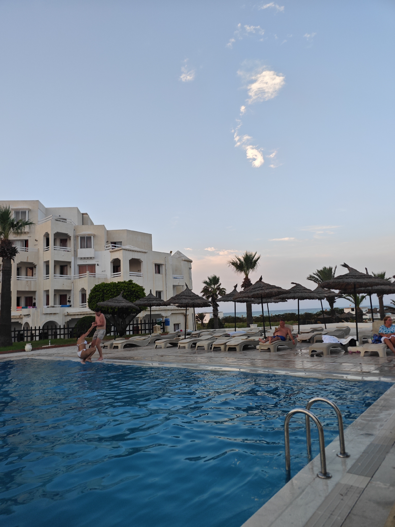 The image shows a resort in Hiboun, Tunisia, on a bright, clear day. In the foreground, a swimming pool with blue water is visible, with metal steps leading into it. Beyond the pool, a beige-colored hotel building with multiple stories and balconies stands under a pale blue sky with scattered white clouds. In front of the hotel, a man and a woman in swimwear are interacting near the edge of the pool. Several rows of empty lounge chairs are arranged on a paved area, some covered by straw umbrellas, and palm trees are scattered throughout the scene. The overall atmosphere suggests a holiday resort setting.