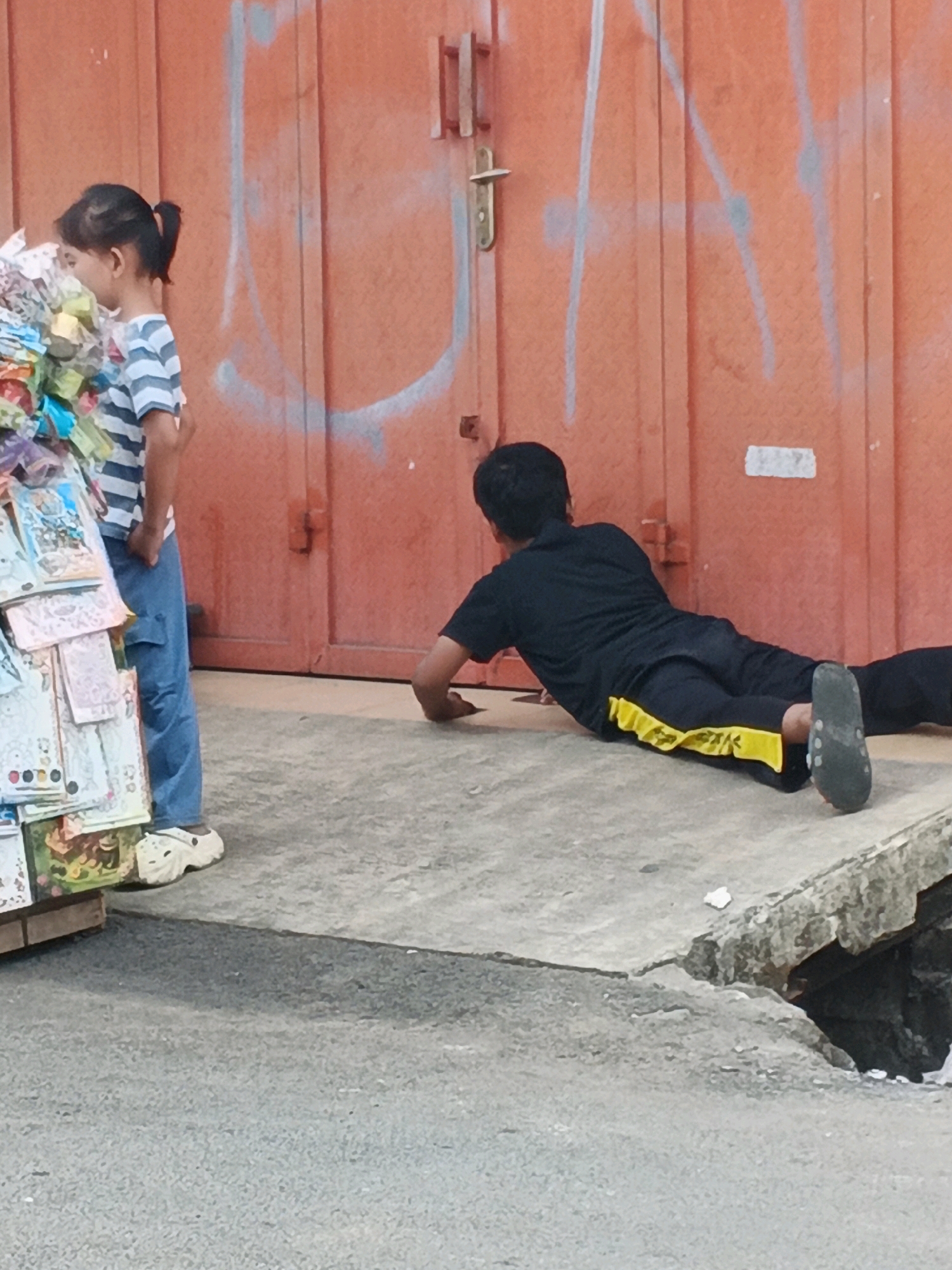 In Kadu, Indonesia, a young girl stands to the left of the frame, facing away from the viewer, examining a display of assorted merchandise. She is wearing a striped t-shirt and blue pants. To the right, a person wearing a black t-shirt and black pants with a yellow stripe is lying on the ground. The person appears to be either resting or performing an exercise on the concrete surface. The background consists of a large, orange, corrugated metal door with some graffiti. The scene takes place outdoors on a sidewalk or street.