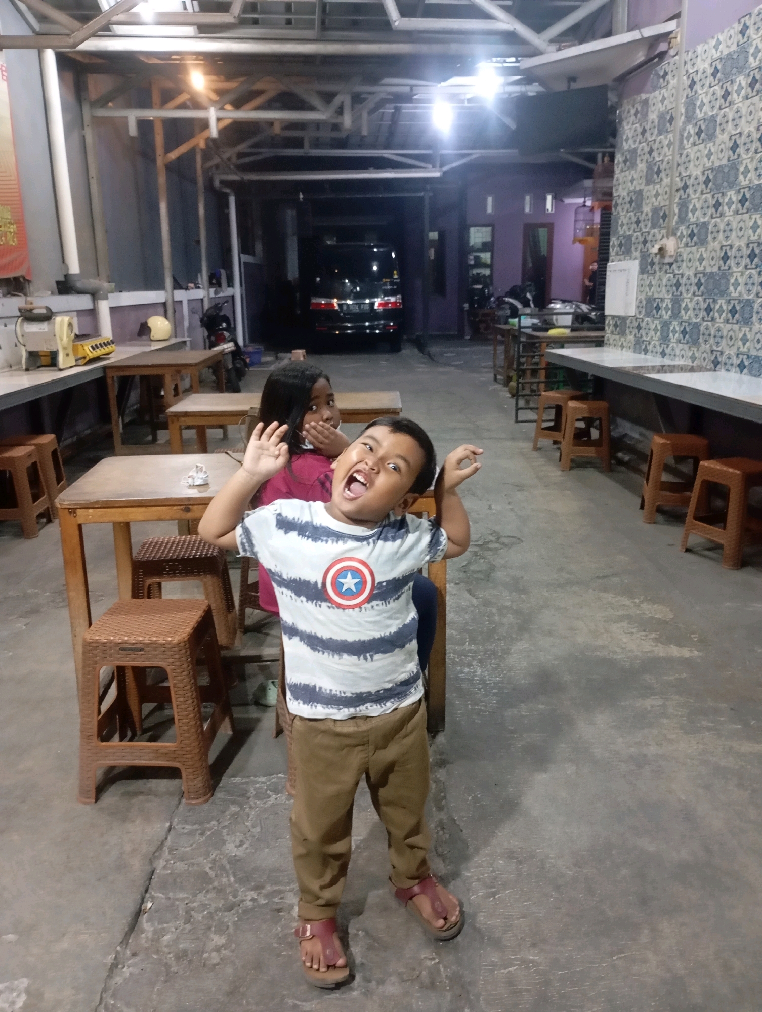 The image depicts an outdoor seating area of a food establishment, likely in Babelan, Indonesia, with a concrete floor and a covered ceiling supported by metal beams. In the foreground, a young boy with a wide, open mouth and raised hands stands facing the camera. He is wearing a striped t-shirt with a Captain America emblem and brown pants. To his left and slightly behind him, a young girl with dark hair covers her mouth with her hand and looks towards the camera. She is wearing a pink shirt. Scattered throughout the scene are wooden tables and brown plastic stools. In the background, a dark-colored van is parked, and part of a building with patterned tiles is visible. The lighting suggests it is evening or dusk.