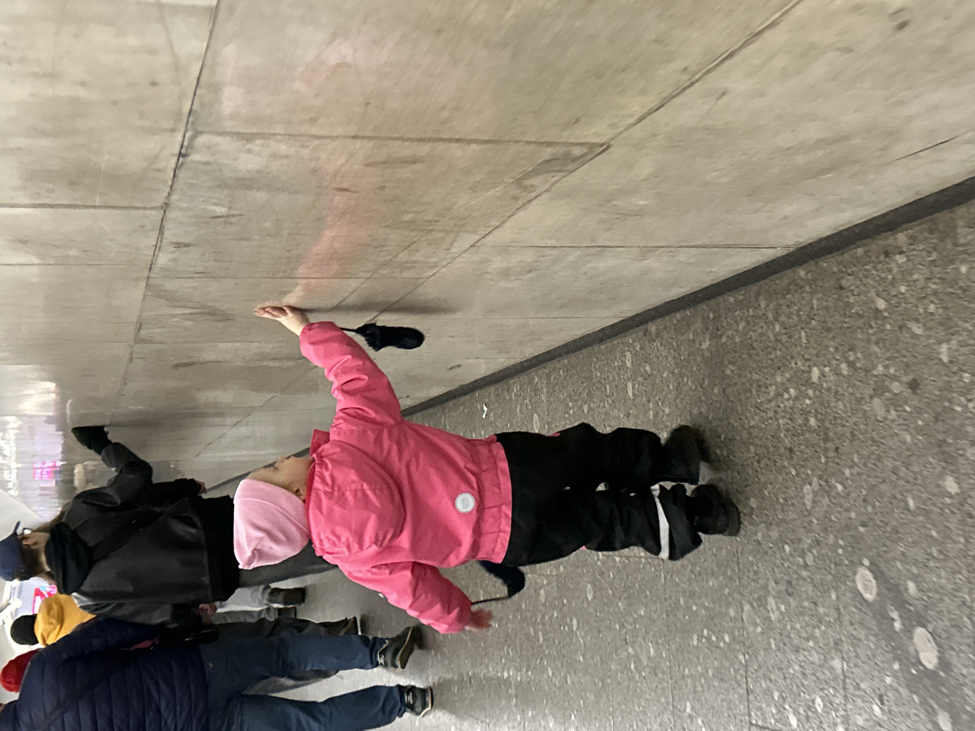 This image depicts a scene inside an indoor or covered public space, likely an underpass or a public transport station in Saint Petersburg, Russia. The perspective is a slight low-angle shot, looking up towards a group of people and the architectural elements of the structure.
In the foreground, a young child is visible from behind, dressed in a bright pink puffer jacket, a light pink beanie, and black winter pants with white reflective stripes near the ankles. The child's right arm is raised, pointing or reaching towards the light grey paneled ceiling or upper wall, which is constructed from large rectangular concrete sections with visible dark seams. A small, dark object, possibly a folded umbrella or a bag strap, hangs near the child's outstretched hand, casting a faint shadow.
Behind the child, an adult or older youth is seen wearing a dark jacket and a dark baseball cap. Further back, another person in a bright yellow jacket is partially visible, and a fourth individual in a dark blue jacket and blue jeans can be seen further in the background. The people appear to be either walking or standing in transit.
The floor is a speckled grey material, resembling terrazzo or polished concrete, with some lighter marks. The overall environment is utilitarian, characterized by dominant grey concrete surfaces. The lighting suggests an artificial indoor setting. No specific activity or event is clearly depicted beyond general movement in a public area, and no emotions are discernible from the visible faces. There is no visible text in the image.