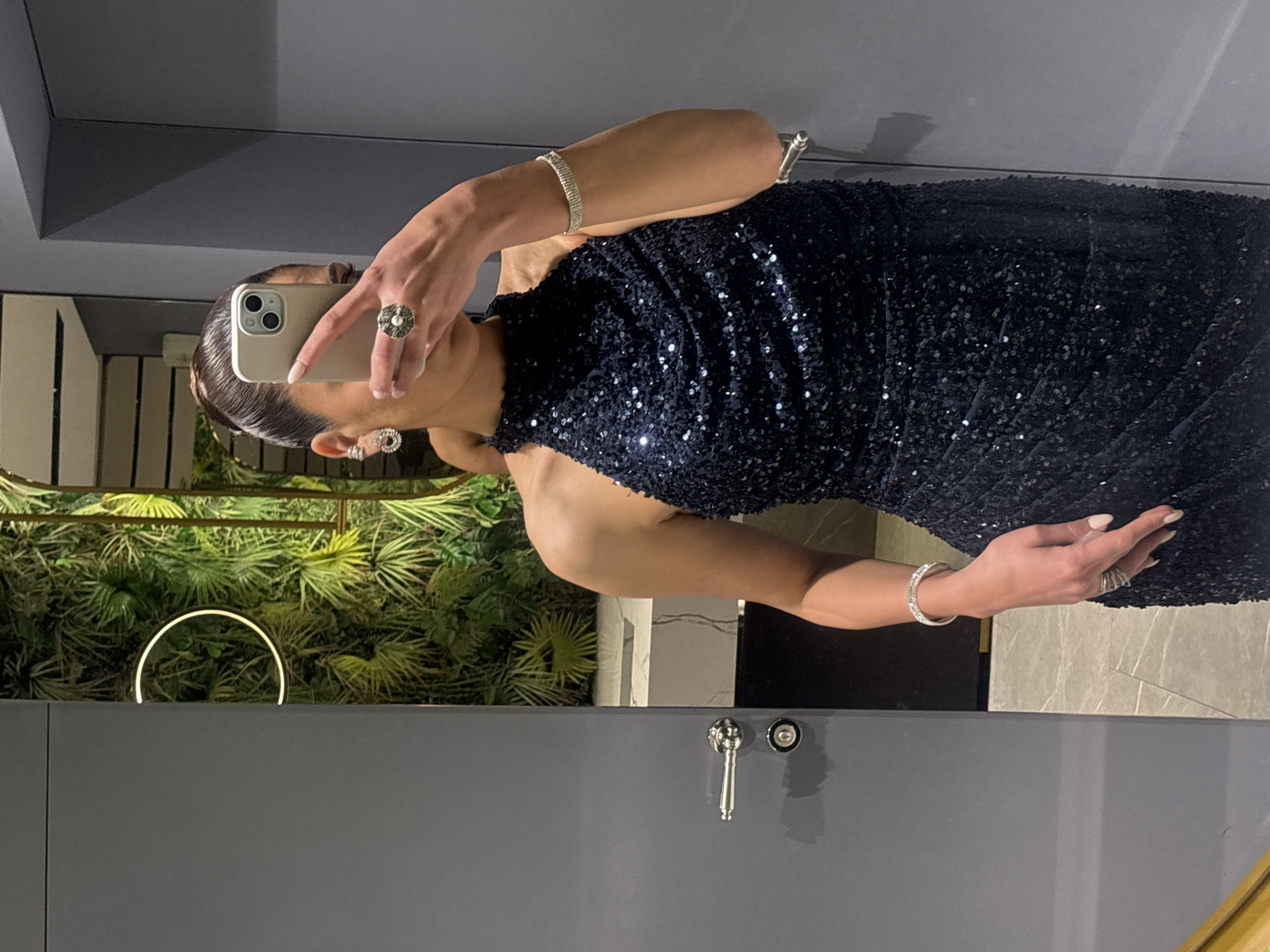A person stands in a bathroom, taking a selfie in a mirror. They are wearing a sleeveless, sequined dress. The person is holding a white smartphone with the camera visible, obscuring their face. On their left wrist is a bracelet, and a ring on their finger. The bathroom has a green decorative wall with a circular light. A tap and sink fixture are visible. The location is presumed to be in Masis, Armenia.