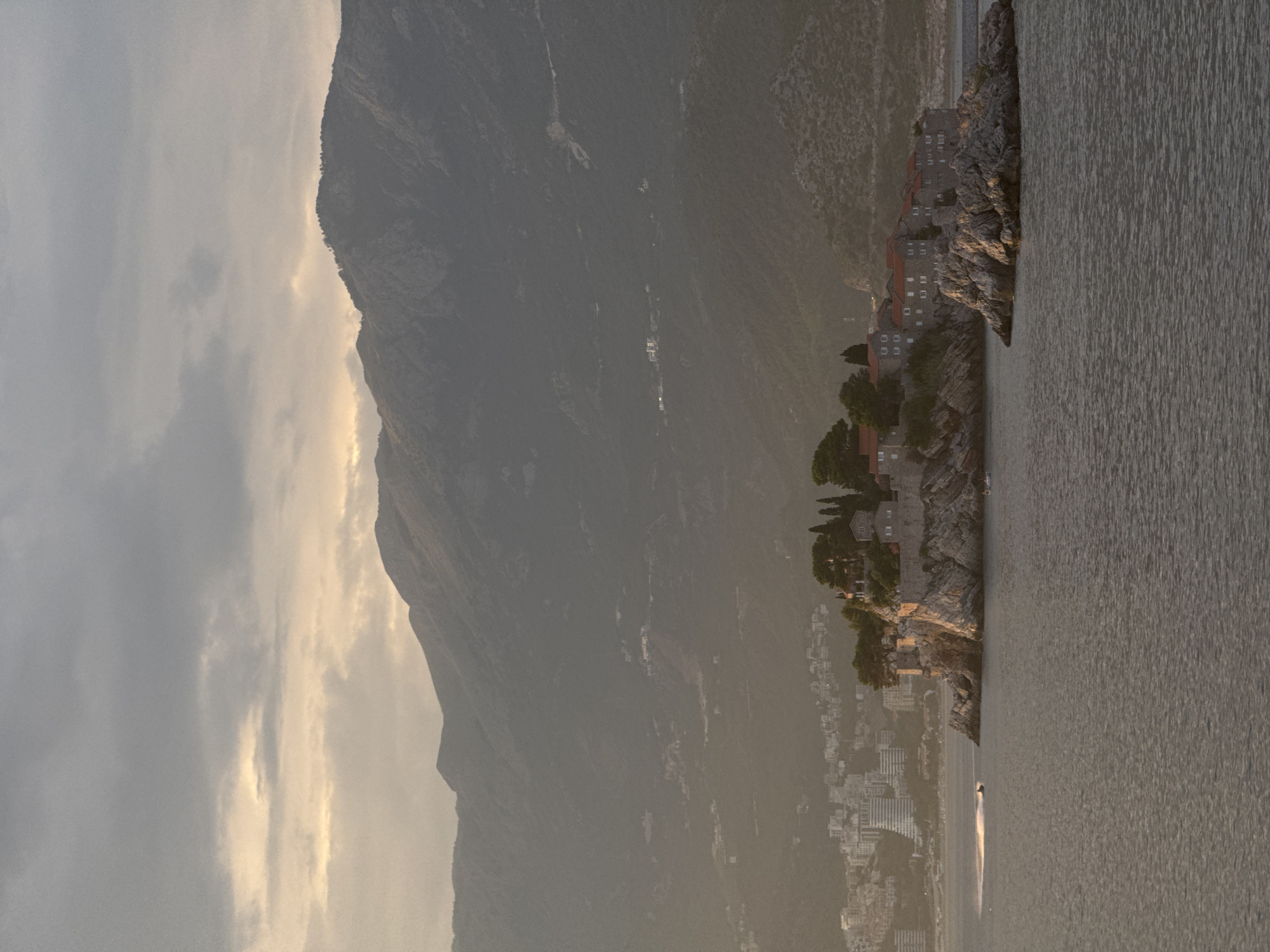 The image shows a view of the island of Sveti Stefan in Montenegro. The island is a small, rocky outcrop connected to the mainland by a narrow causeway. On the island, there are several stone buildings with red tile roofs, surrounded by trees. The buildings appear to be an ancient village. In the foreground, the sea is calm, with a slight ripple. A jet ski is visible in the water, creating a spray of water behind it. In the background, a coastal city with modern buildings can be seen, and behind that, mountains rise into the sky, partially obscured by haze. The sky is cloudy with patches of sunlight breaking through, suggesting either sunrise or sunset. The overall atmosphere is serene and picturesque.