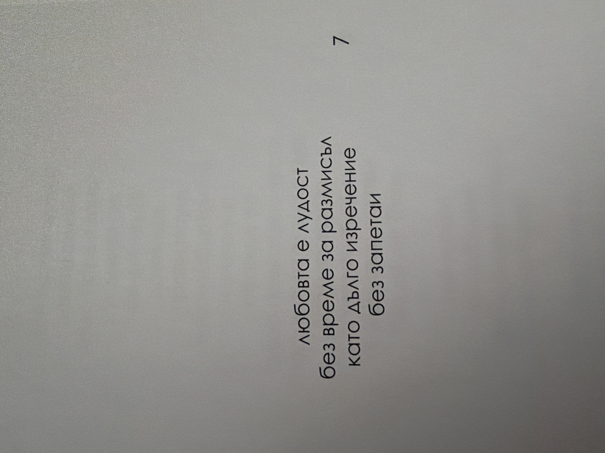 The image is a close-up of a page from a book or document. The background is a plain, light gray, with a subtle textured pattern. Centered on the page are four lines of text in Bulgarian, in a serif font. The text reads:
"любовта е лудост
без време за размисъл
като дълго изречение
без запетаи"
To the right of the text, aligned with the bottom line of text, is the number "7" in the same font. The image does not contain any information that would allow for its location to be identified within the city of Pazardzhik, Bulgaria, or any other specific location.