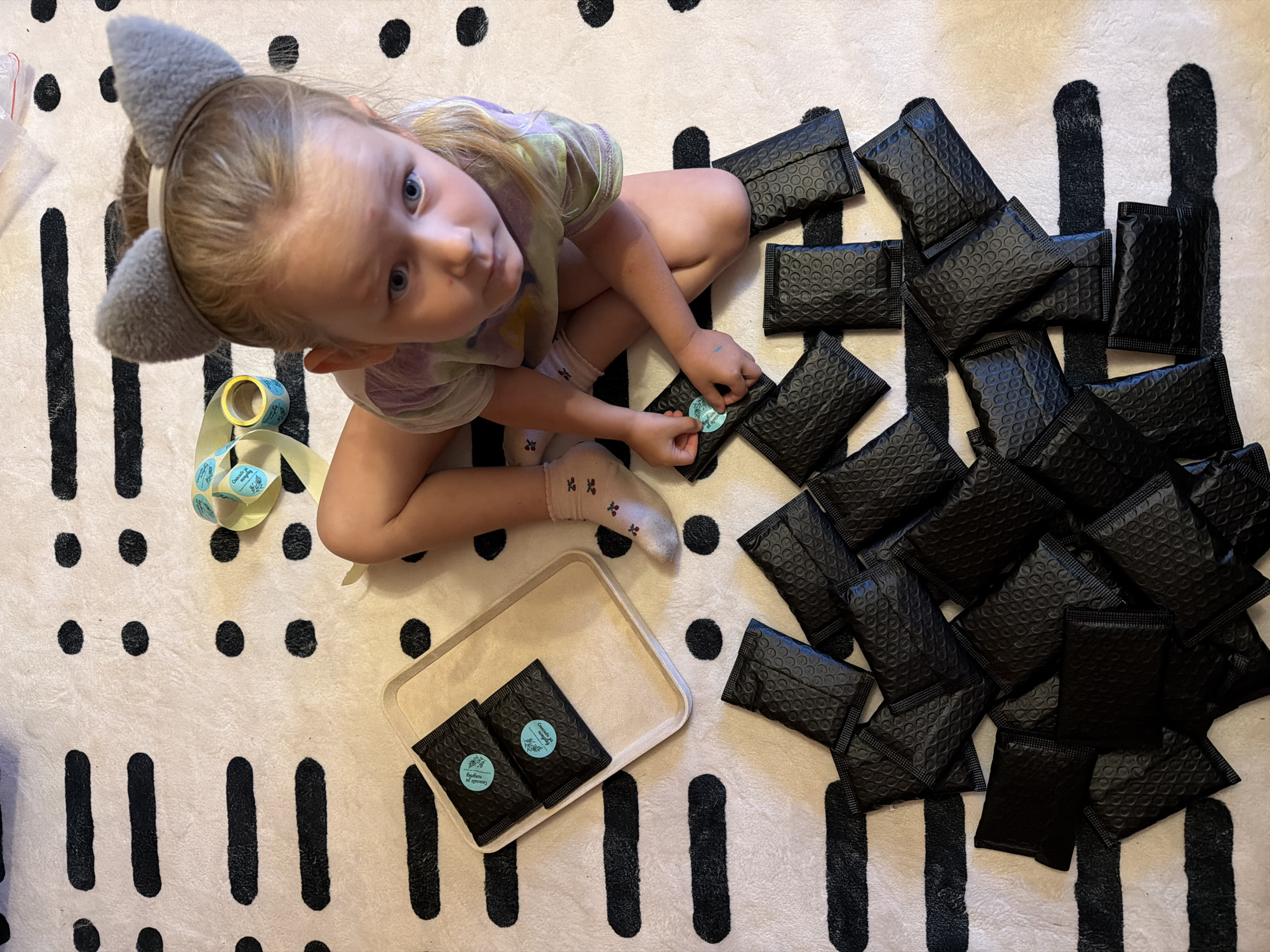 A young child, wearing grey cat ears and a colorful t-shirt, sits on a rug with a black and white striped and polka-dotted pattern. The child is surrounded by numerous small black padded envelopes, most of which are arranged in a scattered pile. Two of these envelopes are placed in a light-colored rectangular tray to the child's left. A roll of yellow tape with blue circular stickers is also visible near the child. The child is looking up at the camera with a curious expression. One of the stickers on an envelope reads "Cascade Oil & Beauty". The lighting suggests it is daytime. The overall scene appears to be indoors, possibly in a child's room or play area.