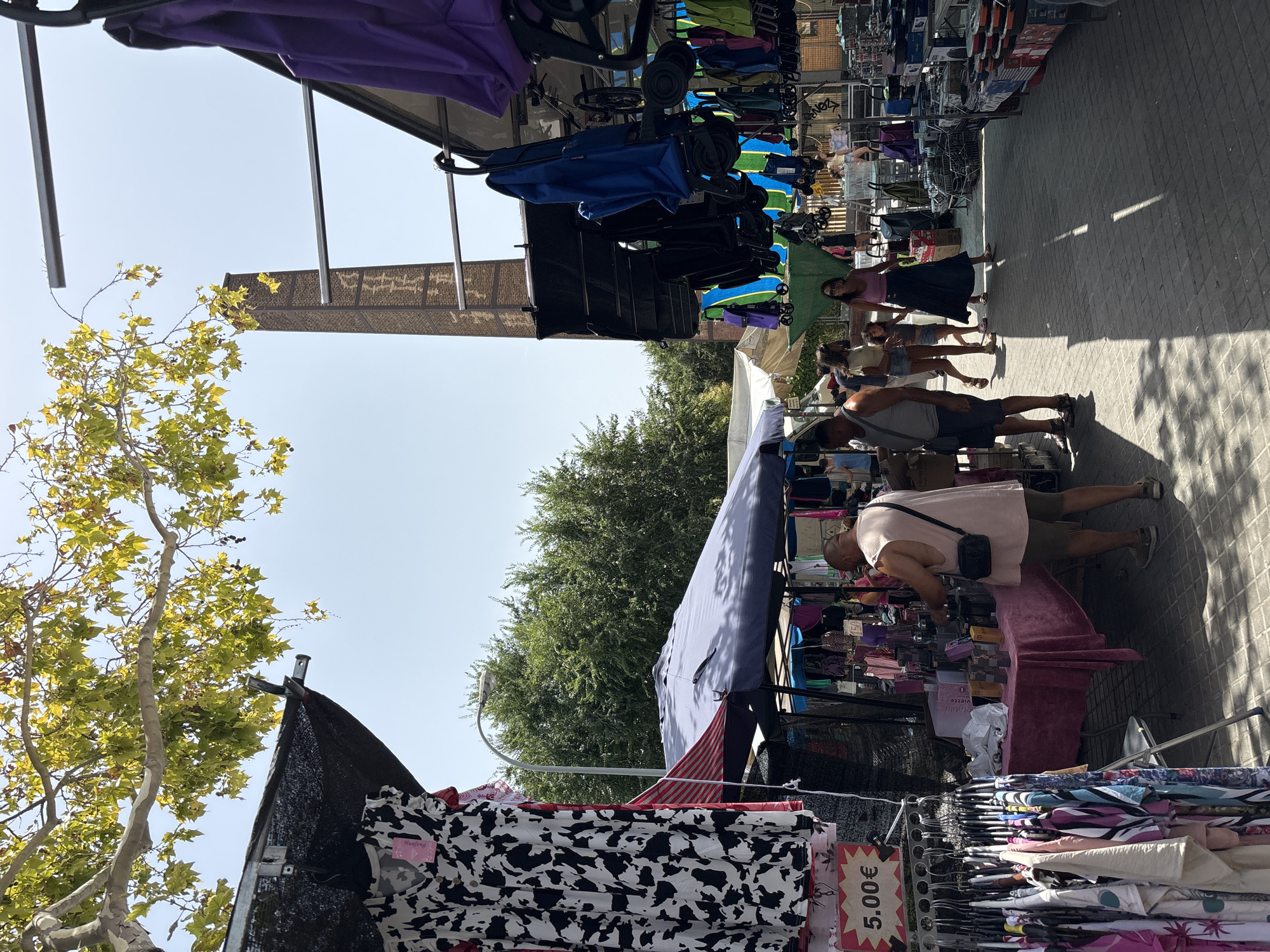 The image depicts an outdoor market scene, possibly in Badalona, Spain, characterized by stalls selling clothing and other merchandise. In the foreground, racks of colorful clothing are displayed. A price tag reading "5.00€" is visible on one of the garments. A man in a light-colored sleeveless shirt and shorts is browsing items at a stall covered by a blue and red awning. He is wearing a dark cross-body bag. Several other people are walking through the market. In the background, a tall brick chimney with architectural details stands against a clear sky. Trees with green and yellow leaves are visible overhead, and a street lamp is mounted on a curved pole. The ground is paved with grey interlocking bricks, and shadows from the stalls and trees are cast across the walkway.