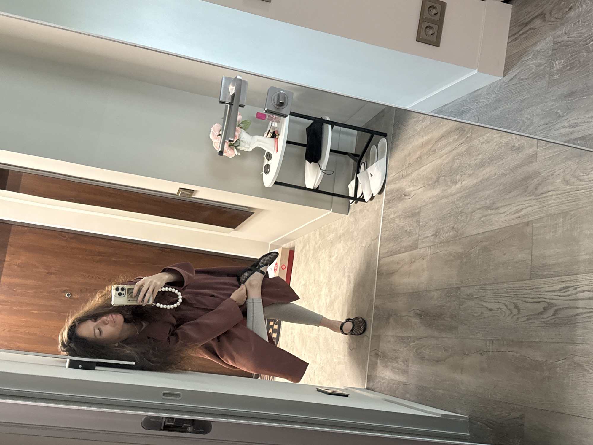A woman is captured in a mirror selfie taken in what appears to be a modern home entryway or hallway. The woman, with long, wavy dark hair, is reflected holding a light-colored smartphone (possibly an iPhone) with a distinctive white beaded wrist strap. She is dressed in a long, open burgundy or maroon coat over light grey leggings and black mesh flat shoes with studded details and ankle straps. Her expression is neutral to slightly pouting as she looks towards her phone's camera, with one leg bent and appearing to adjust her shoe.
The background, visible in the mirror's reflection, features a dark wooden door to her right. To her left, against a light grey wall, there's a multi-tiered white and black metal storage shelf. The shelf holds various items including a vase with pink artificial flowers, a small bottle (possibly perfume), sunglasses, black fabric (likely a mask or small pouch), and two pairs of white slippers. Above the shelf, a light fixture and a double electrical outlet are mounted on the wall. The flooring throughout the area is composed of light grey wooden laminate panels. A small dark object is also visible on the floor near her feet. The scene suggests an indoor environment, likely a well-lit daytime setting, in Moscow, Russia, as specified by the user. No discernible text is visible in the image.