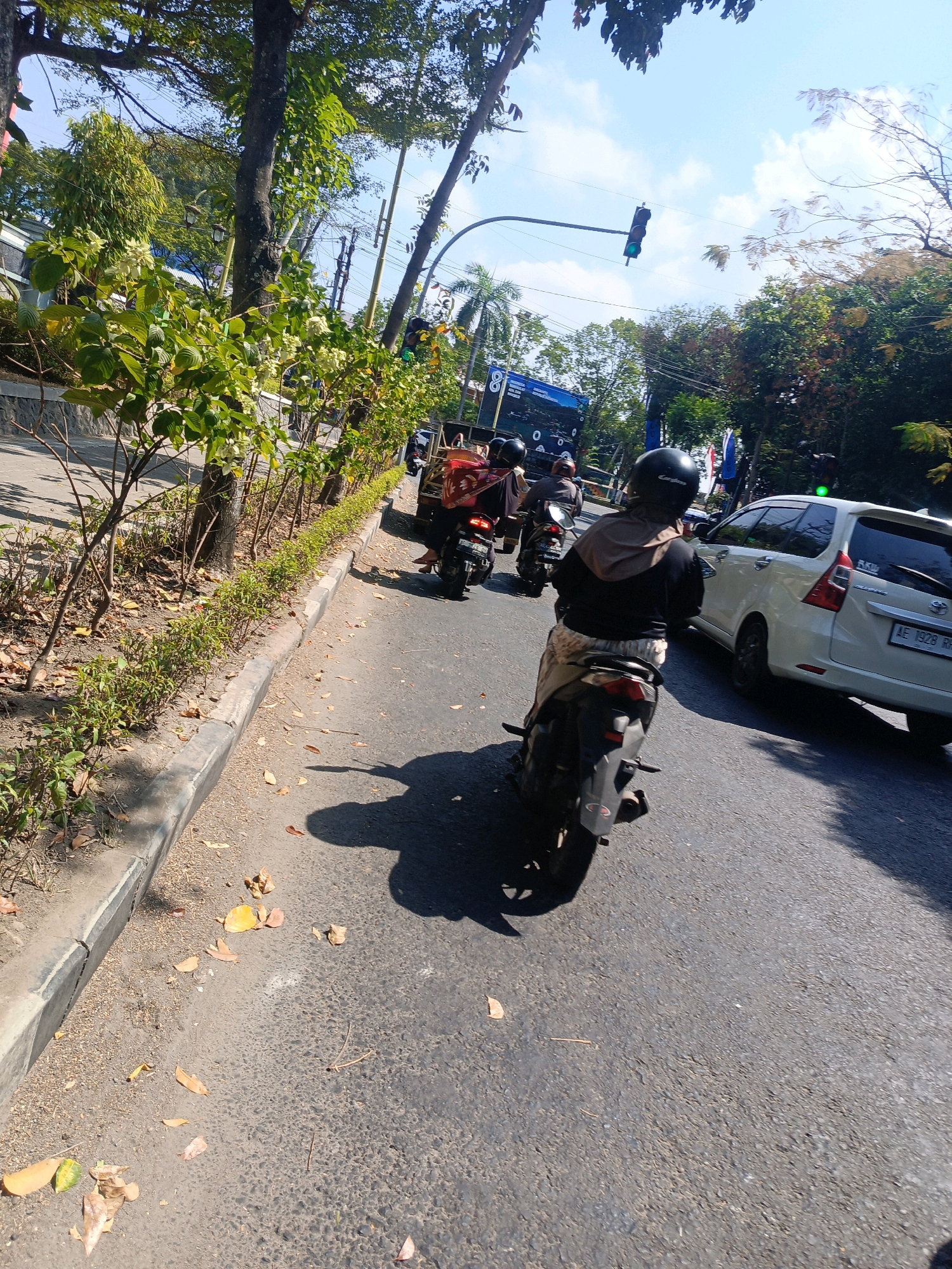 The image depicts a street scene in Tambran, Indonesia, on a bright, sunny day. In the foreground, a person is riding a motorcycle, viewed from behind. They are wearing a black helmet and a light brown headscarf, along with a black hooded jacket and patterned pants. The motorcycle is dark colored, and its rear wheel and fender are visible. The road is paved with asphalt, and scattered dry leaves are present on the surface. To the left of the road, a sidewalk with a concrete curb is lined with small green bushes and several trees. The trees have green foliage, and one is prominent in the mid-left of the frame with its branches extending upwards.
In the background, traffic is visible. Another motorcycle is ahead of the first, with two individuals on it, one wearing a helmet. Further ahead, a white car is in motion, displaying a license plate that reads "AE 1928 RR". A traffic light is suspended over the road, with a green light illuminated. Other trees and foliage line the right side of the road, and in the distance, a glimpse of buildings and potentially other vehicles can be seen. The sky is blue with scattered white clouds.
