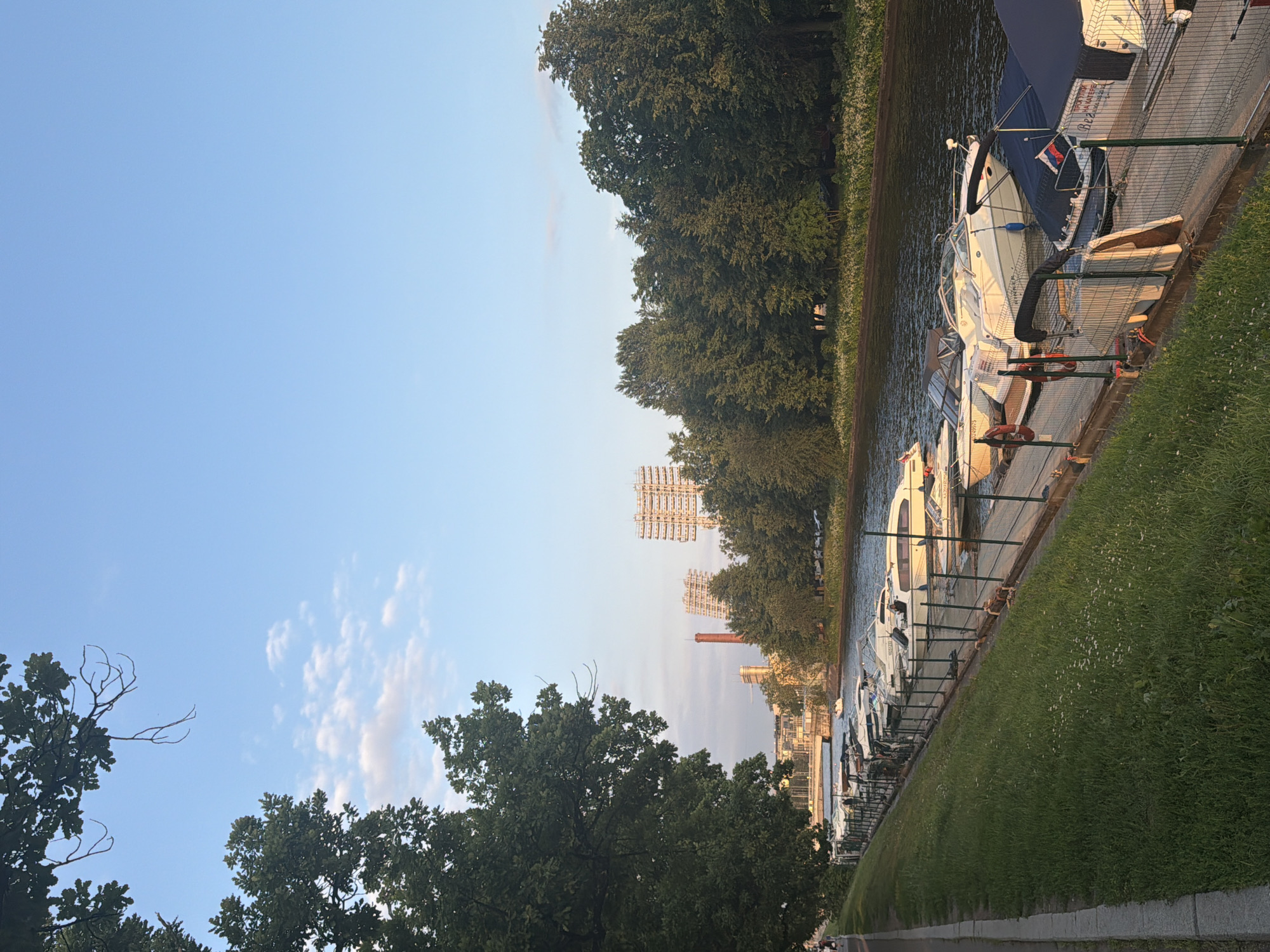 This image depicts a waterfront scene with several boats docked along a pier. The boats are various sizes and types, appearing to be a mix of recreational vessels. The pier is bordered by a grassy embankment on one side and a waterway on the other. In the background, trees line the opposite bank of the water, with some buildings and towers visible in the distance. The sky is clear blue with a few scattered clouds. The sunlight suggests it is either early morning or late afternoon. There is no visible activity of people in the image, but the presence of docked boats implies they are either moored or have recently been used. The overall setting appears to be a calm and serene environment, possibly a marina or a scenic riverside area. There is text visible on the side of one of the boats, but it is too blurry to read.